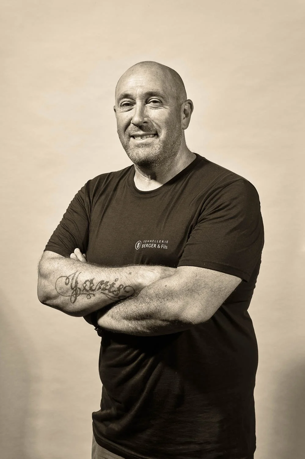 Homme barbu, chauve, souriant, portant un t-shirt noir avec le logo 'TONNELLERIE BERGER & Fils', bras croisés, tatouage sur le bras gauche, fond neutre.