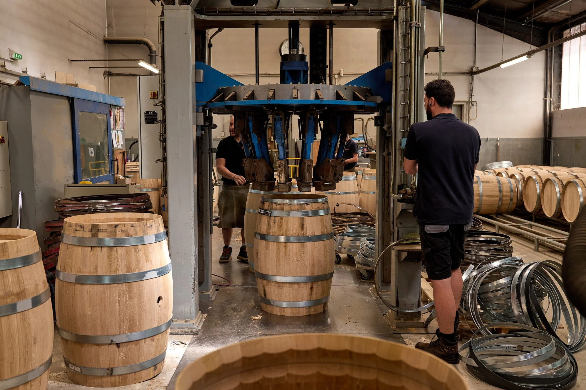 Une scène dans une cave à vin où une machine hydraulique remplit des barriques en bois en utilisant des anneaux métalliques. Plusieurs personnes supervisent le processus, entourées de barriques en bois et de bobines métalliques.