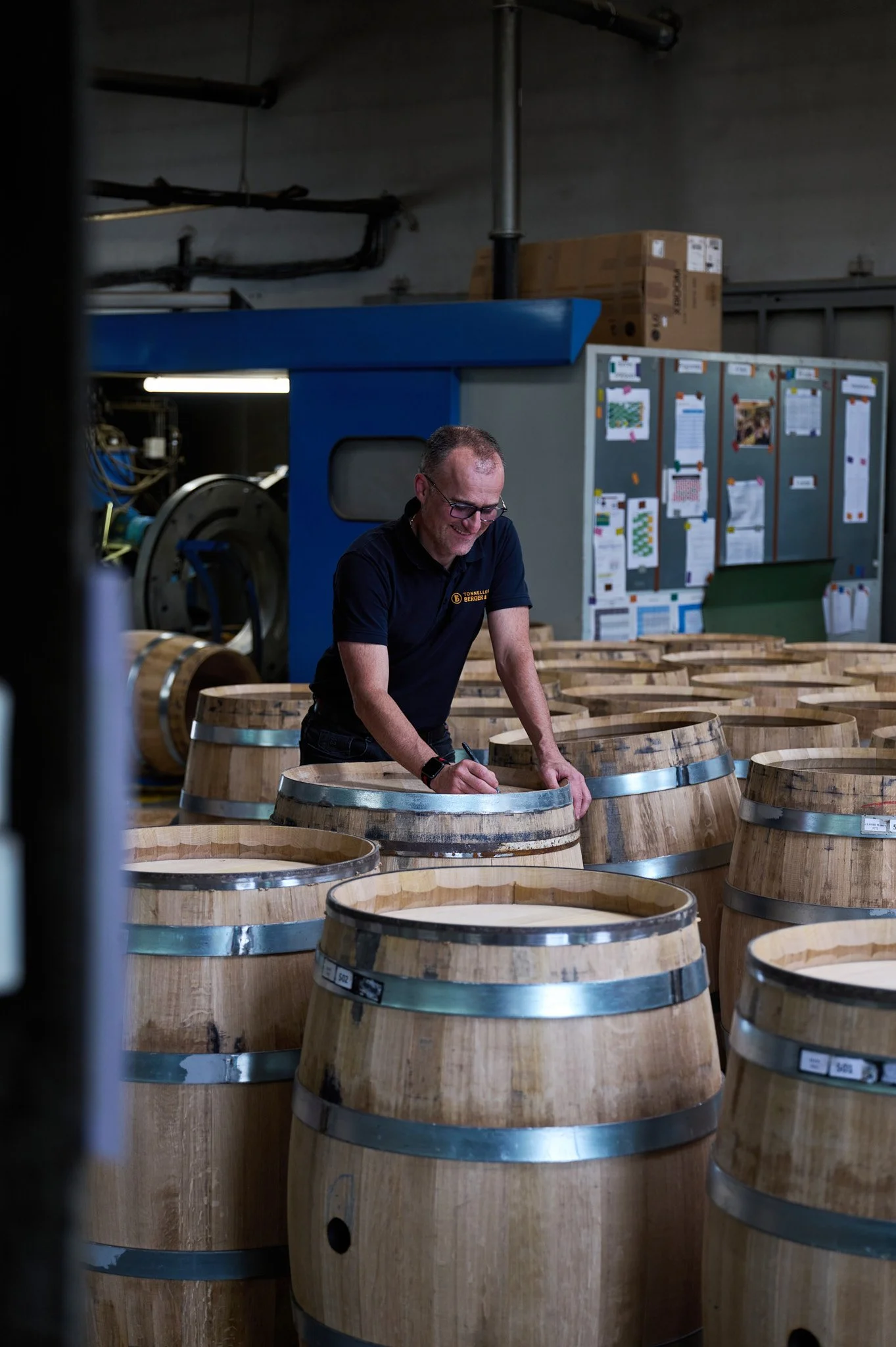 Un homme souriant et portant des lunettes, portant un polo noir, écrit ou signe quelque chose sur un tonneau en bois dans la tonnellerie, entouré de plusieurs autres tonneaux en bois.