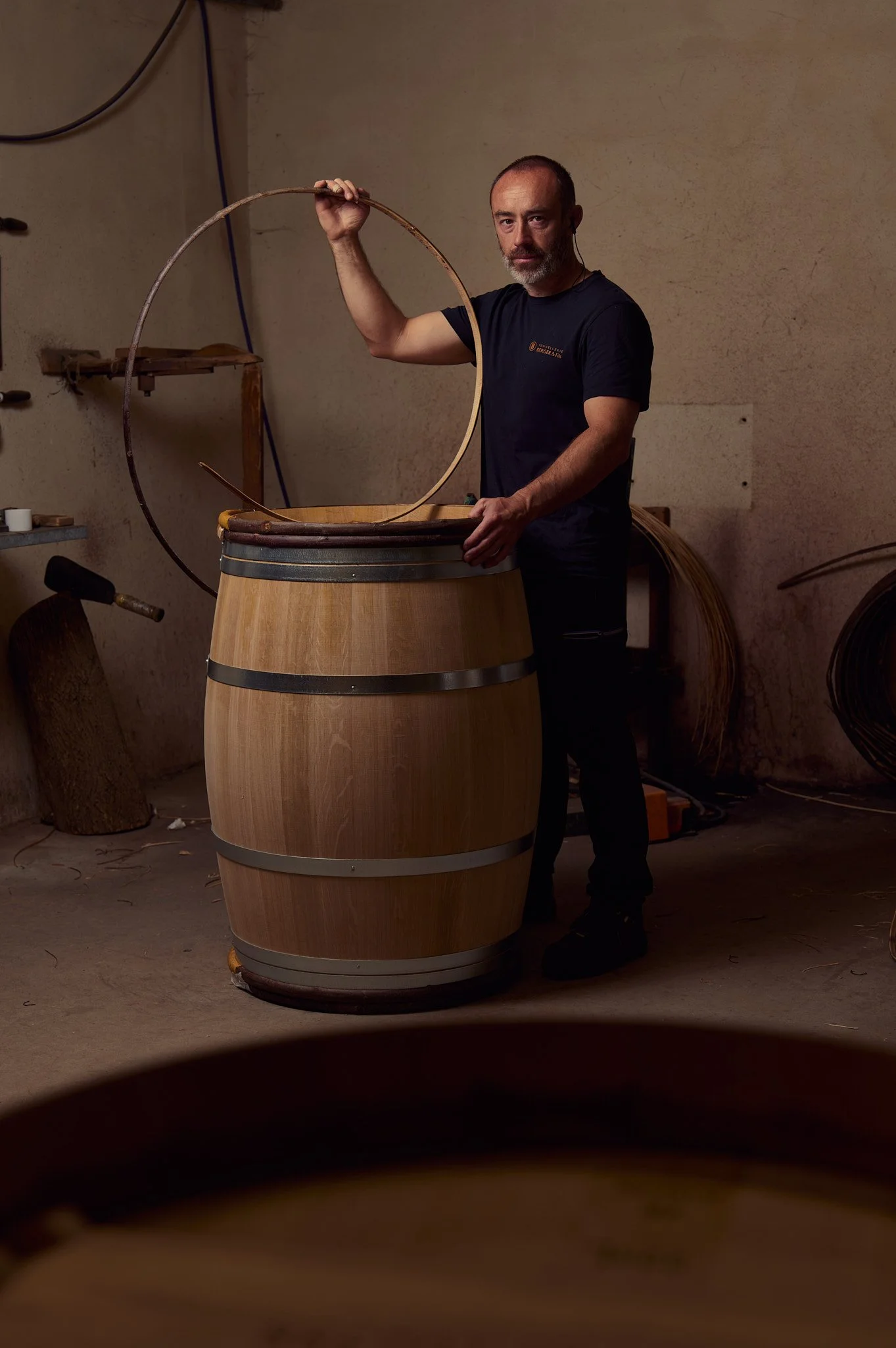 Un homme debout dans un atelier en train de travailler avec du bois, tenant un cercle en bois au-dessus d'une grande cuve en bois, probablement en train de fabriquer un tonneau.