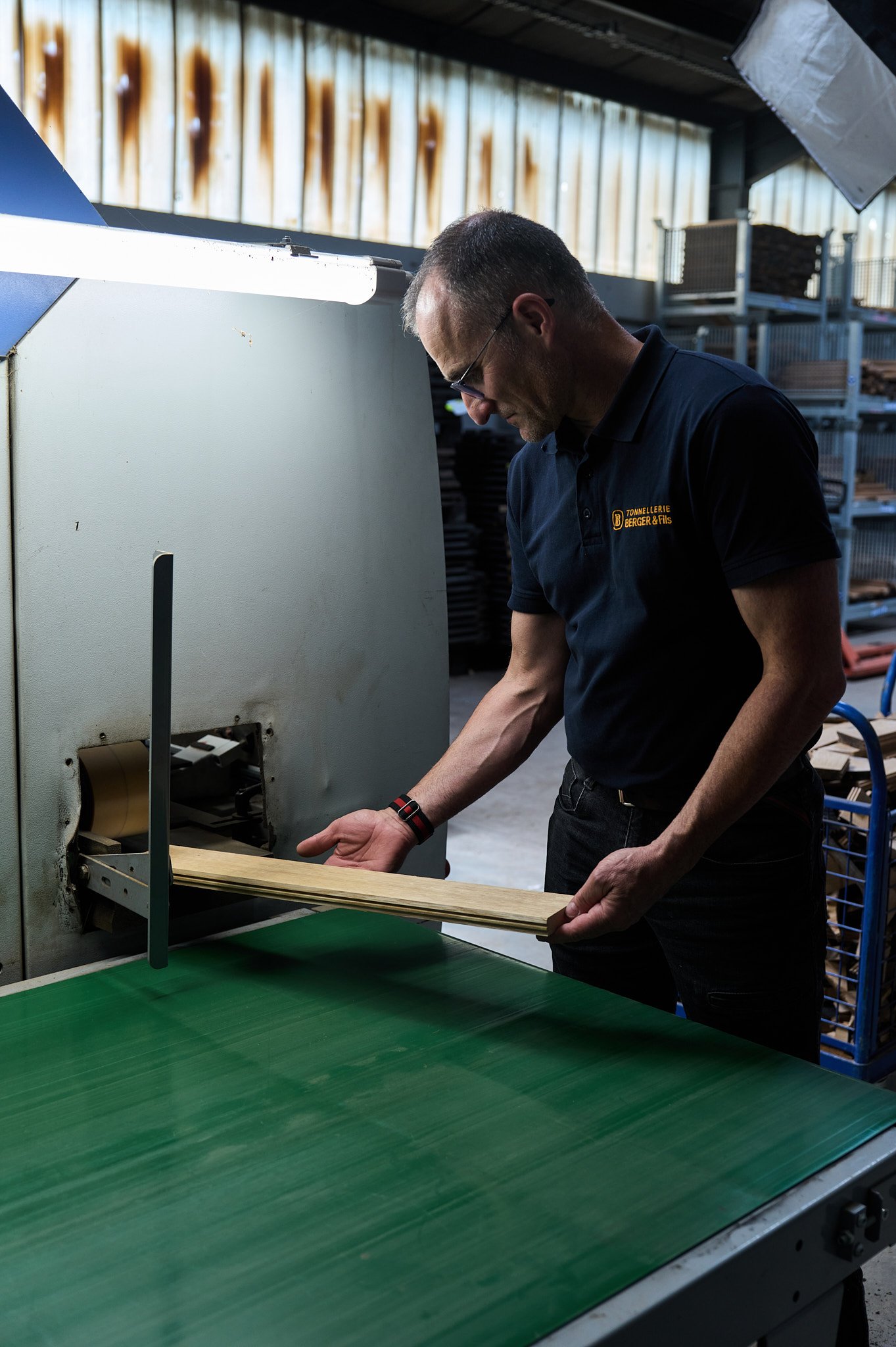 Un homme travaille avec une machine à découper du bois dans une tonnellerie. Il porte un polo noir avec un logo en orange.