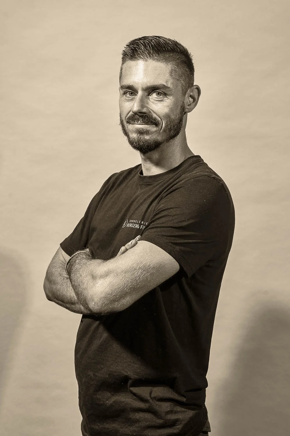 Un homme avec une barbe, portant un t-shirt noir, se tient avec les bras croisés. Il a les cheveux courts et coiffés en arrière, et il sourit légèrement. L'image est en noir et blanc.