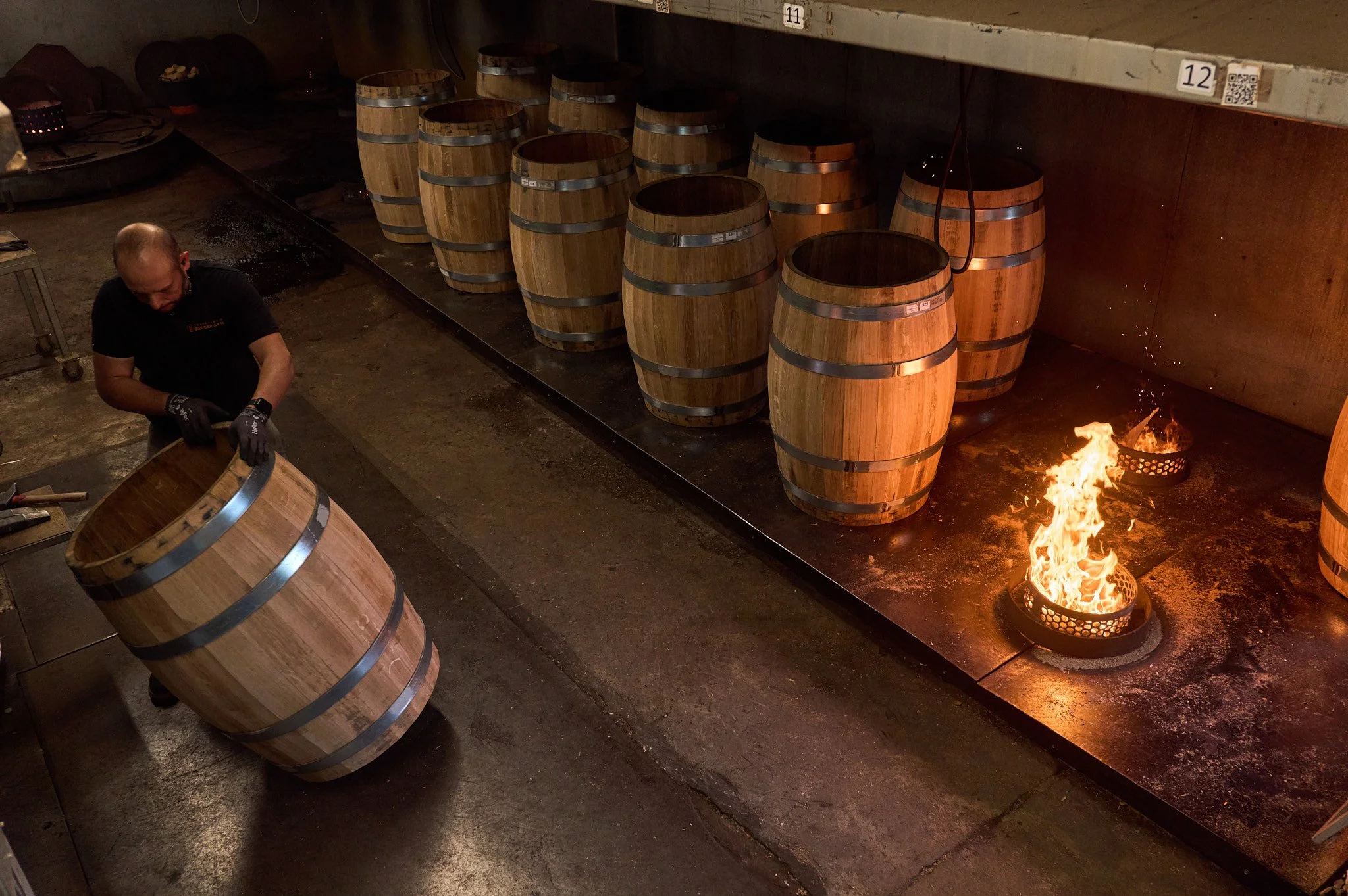 Un artisan tonnelier, manipulant une barrique en bois, avec plusieurs autres tonneaux similaires en arrière-plan et un feu brûlant sur le sol.