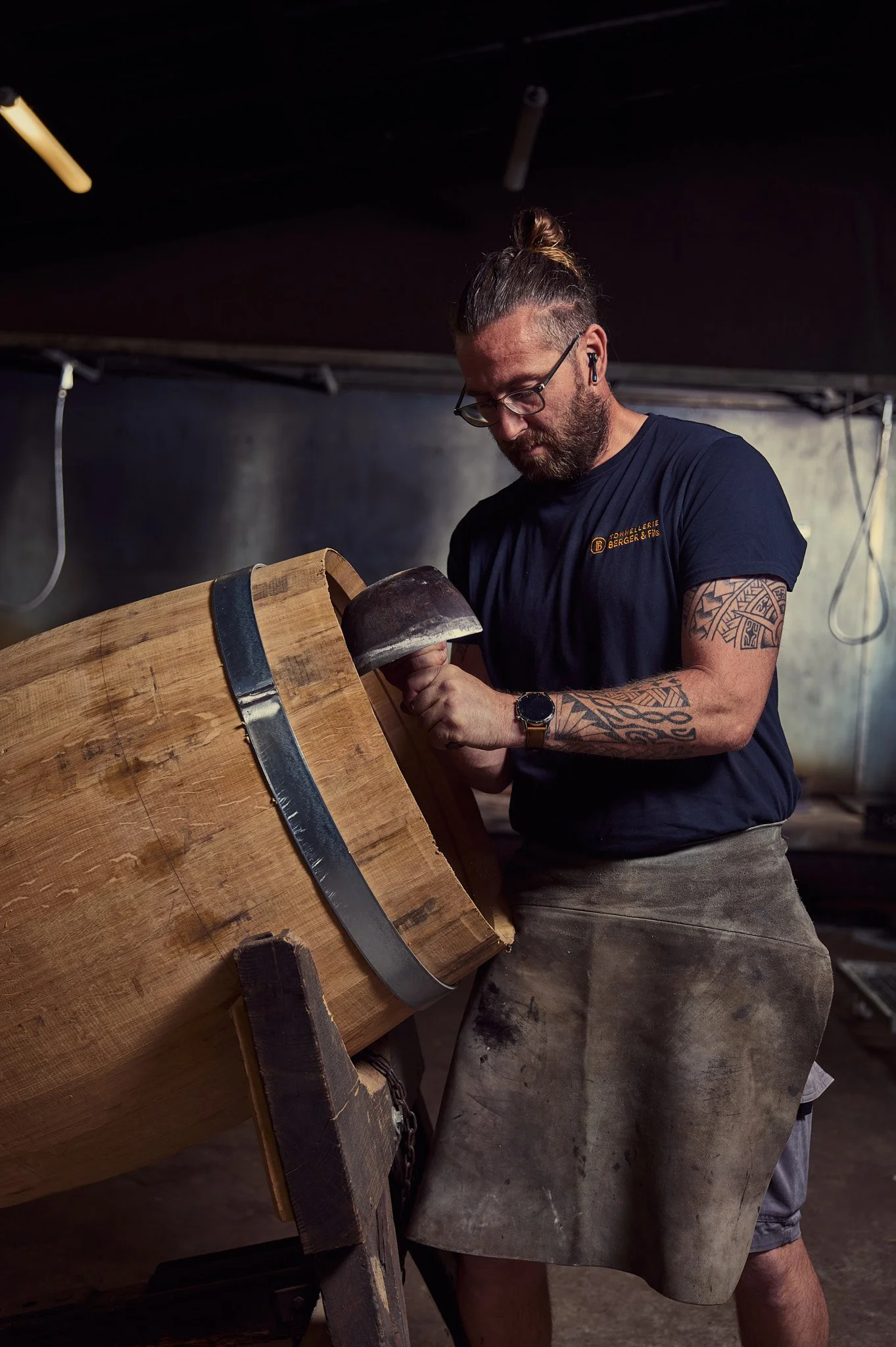 Un homme avec des tatouages, portant une manchette en cuir, travaillant sur un grand morceau de bois dans une forge ou un atelier. Il semble tailler ou façonner le bois en utilisant un outil.