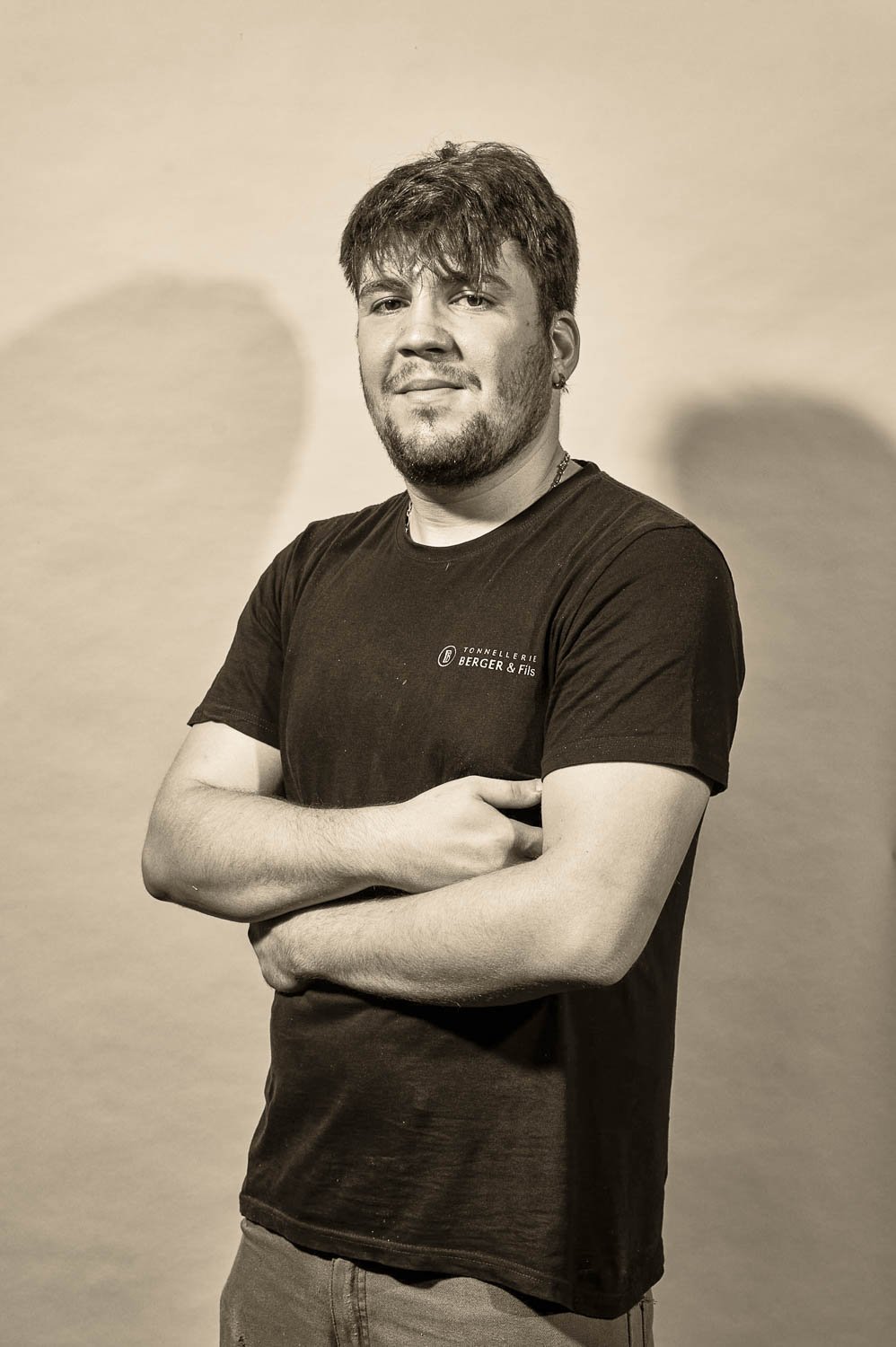 Jeune homme avec barbe et cheveux en désordre, portant un t-shirt noir avec une inscription, les bras croisés, posant contre un mur avec des ombres derrière lui.