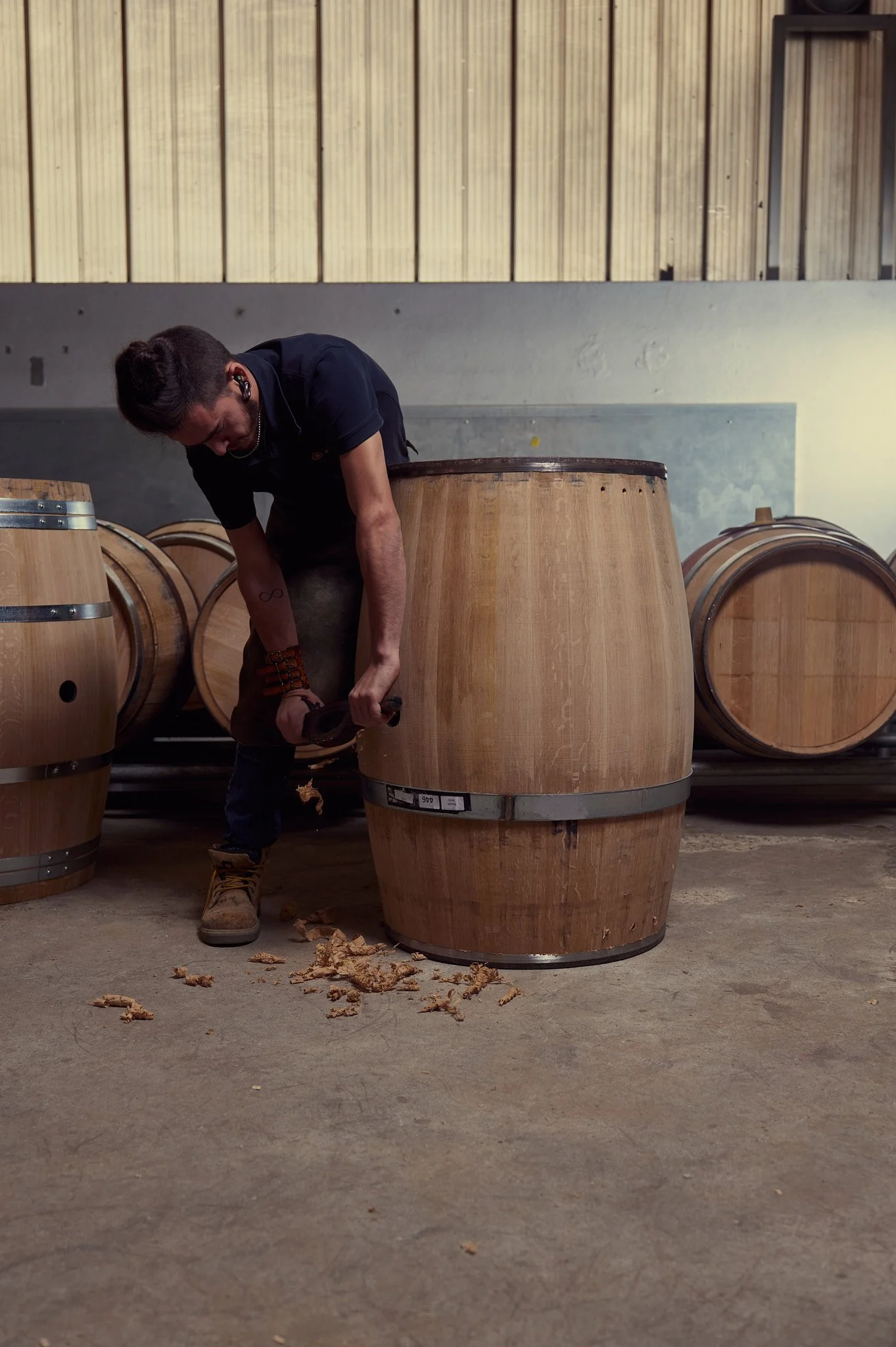 Une personne debout près de barriques en bois dans un atelier, en train de couper ou écailler le bois, avec des copeaux au sol.