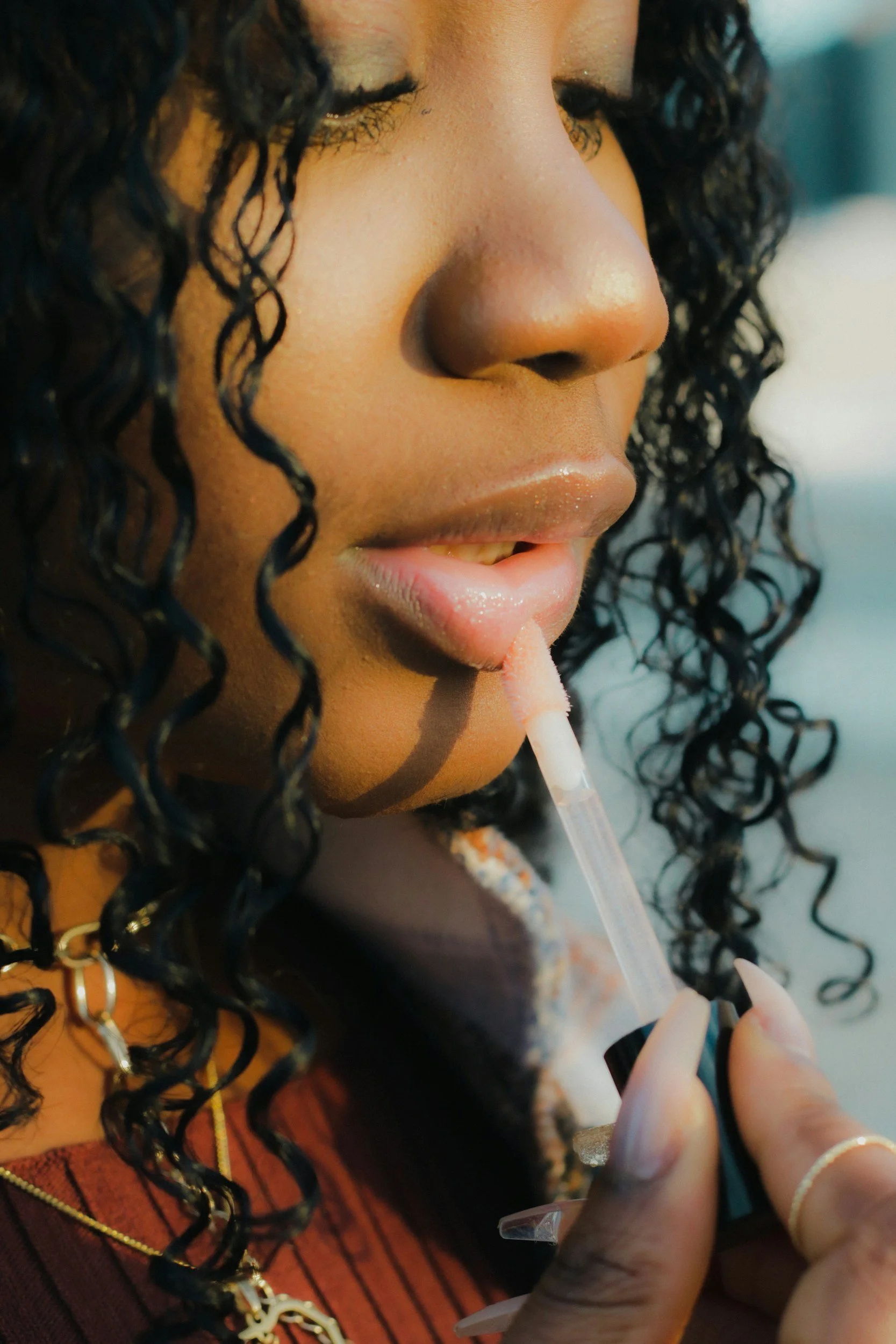 A woman with curly black hair applies gloss to her lips using a lip gloss tube.