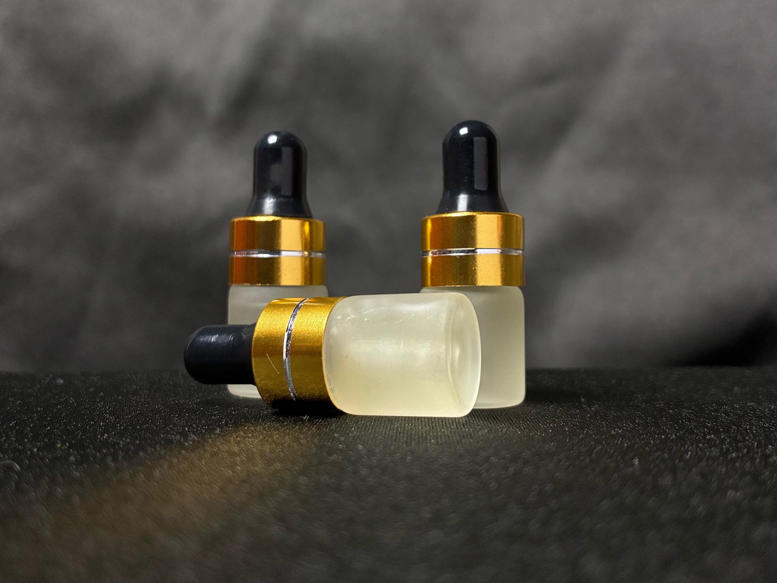 Three small bottles with black dropper caps, gold rings, and frosted glass bodies on a black textured surface with a dark gray background.