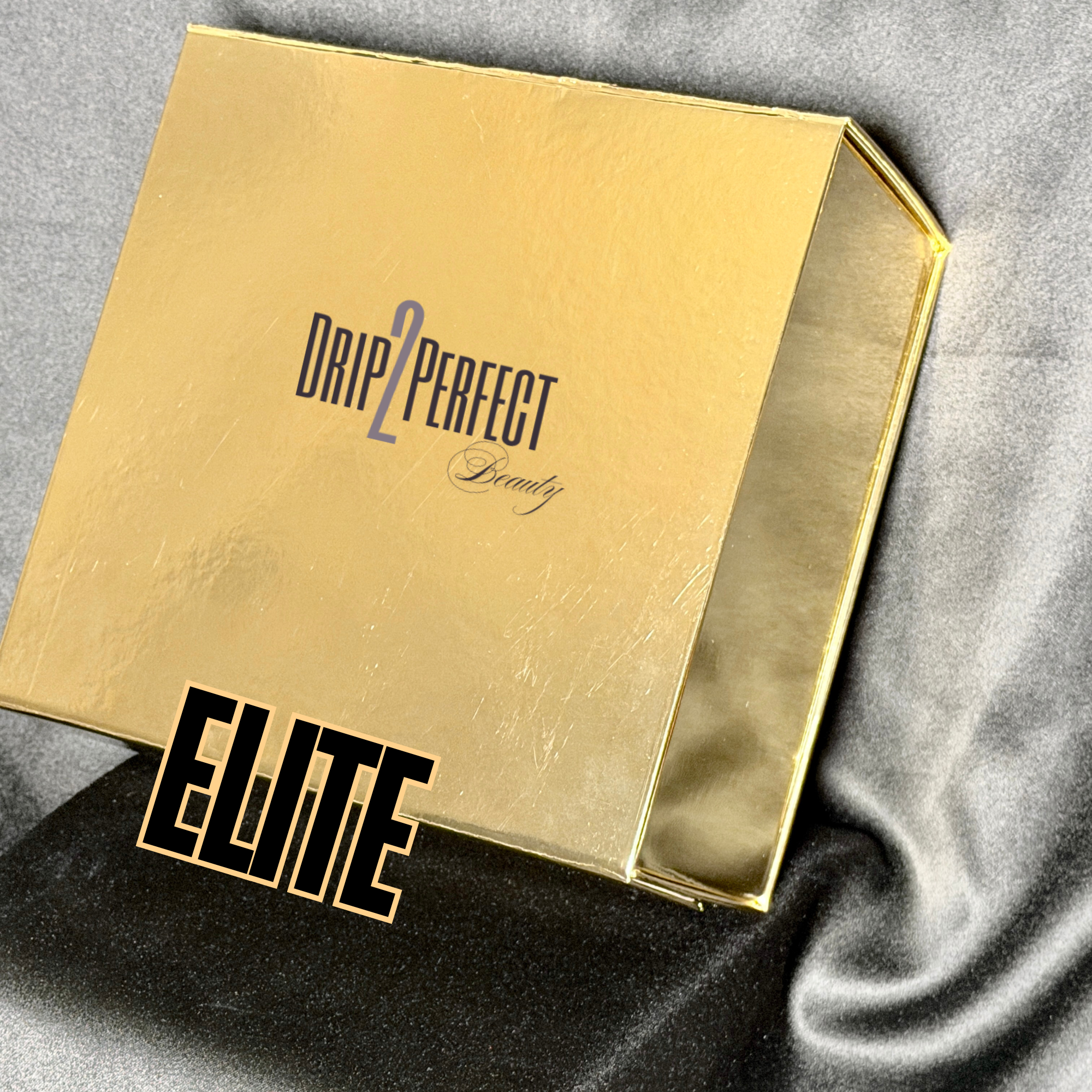 A gold-colored box with the words "DRIP 2 PERFECT Beauty" printed on it, placed on a black satin fabric. The word "ELITE" is overlaid on the image in bold black letters.