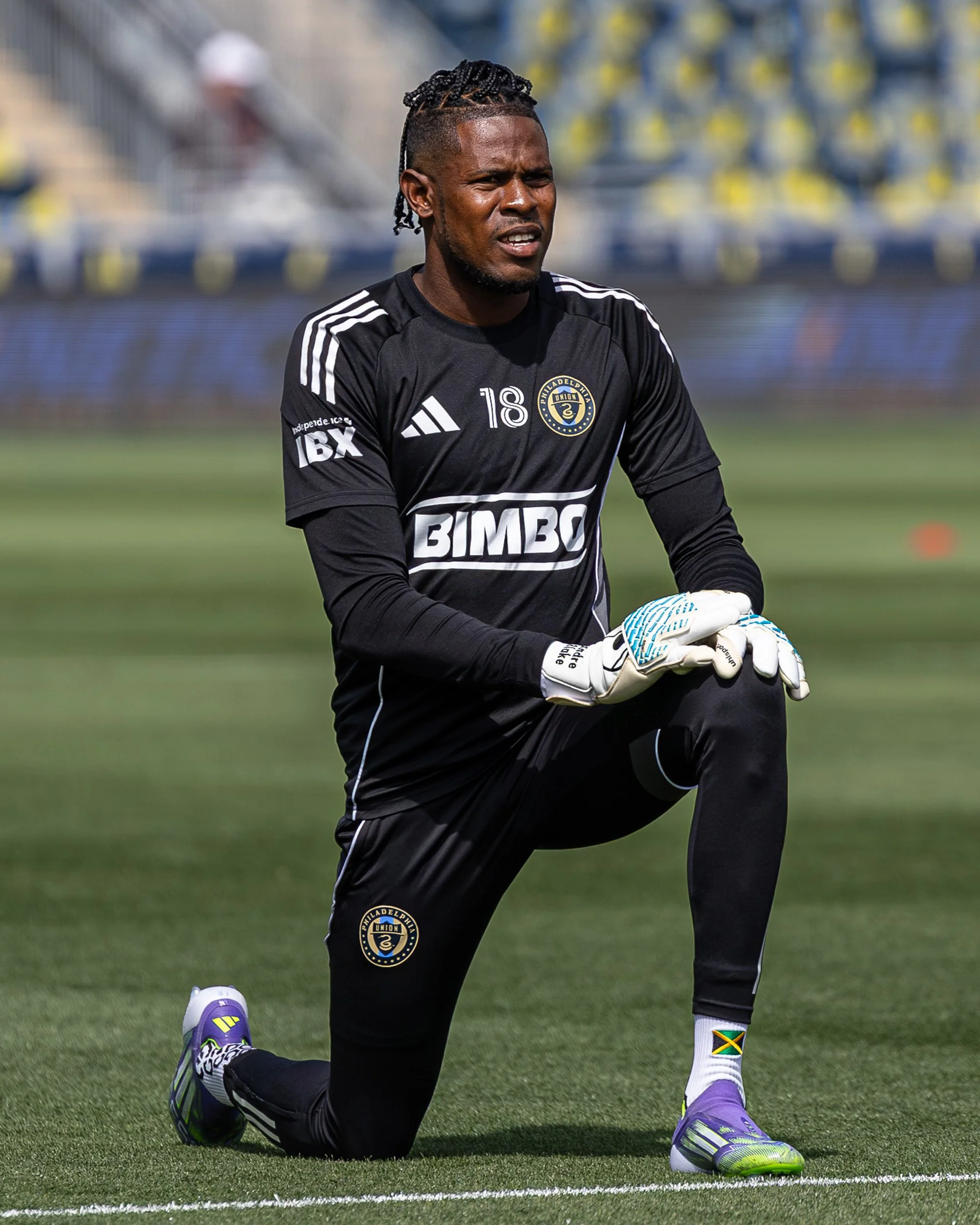 A male soccer goalkeeper kneeling on the field, wearing black uniform and gloves, with a focused expression.