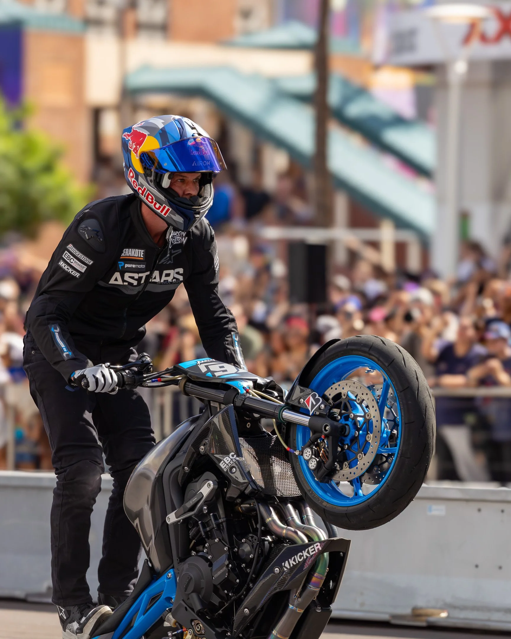 Motorcycle stunt rider performing a wheelie during a public event with a blurred crowd in the background.