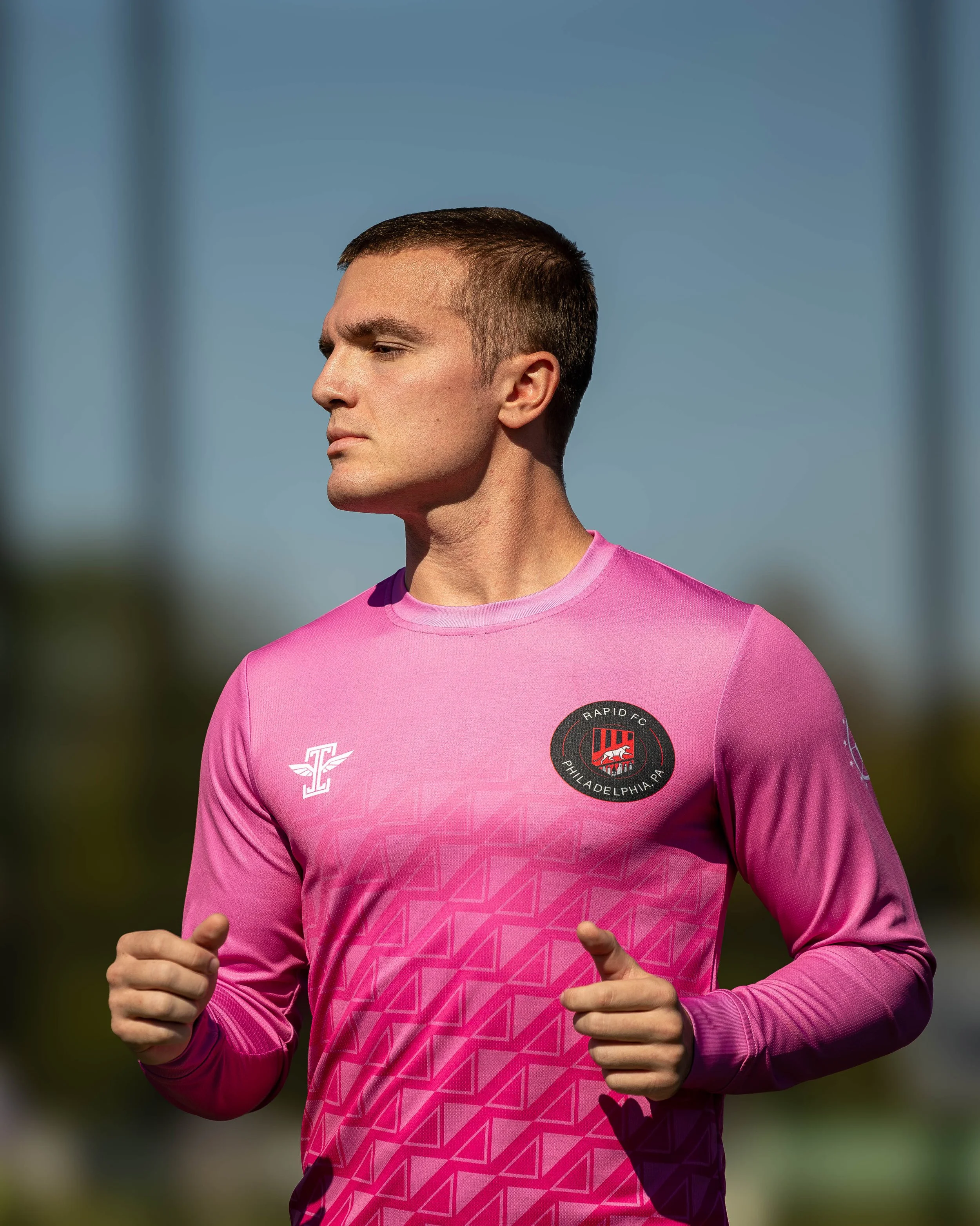 A male soccer player wearing a pink jersey with a Philadelphia Union logo and a white logo on the chest, running outdoors under a blue sky.