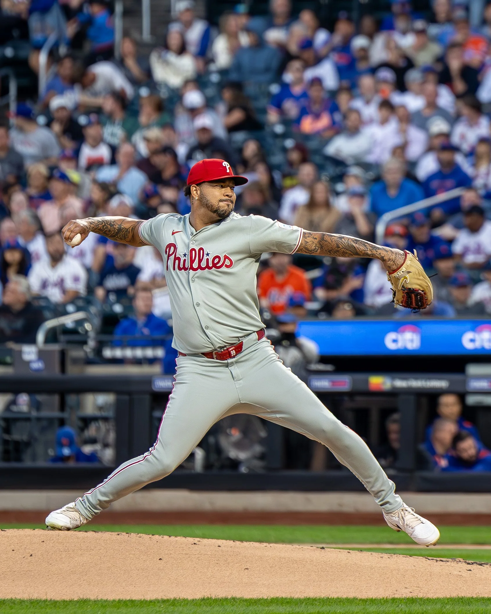 A baseball player from the Philadelphia Phillies in a grey uniform with red accents, wearing a red cap, is pitching on the mound with a crowd of spectators in the background.