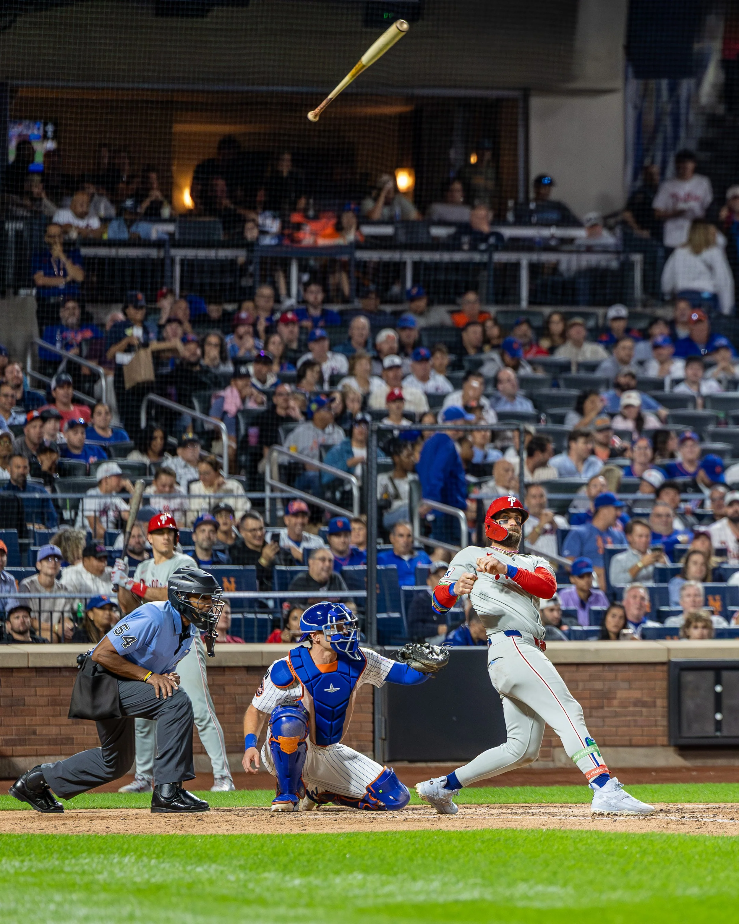 A baseball player in a white uniform and red helmet running after hitting the ball during a game, with a catcher in blue gear, an umpire, and another player behind him on the field, and a large crowd in the stands watching.