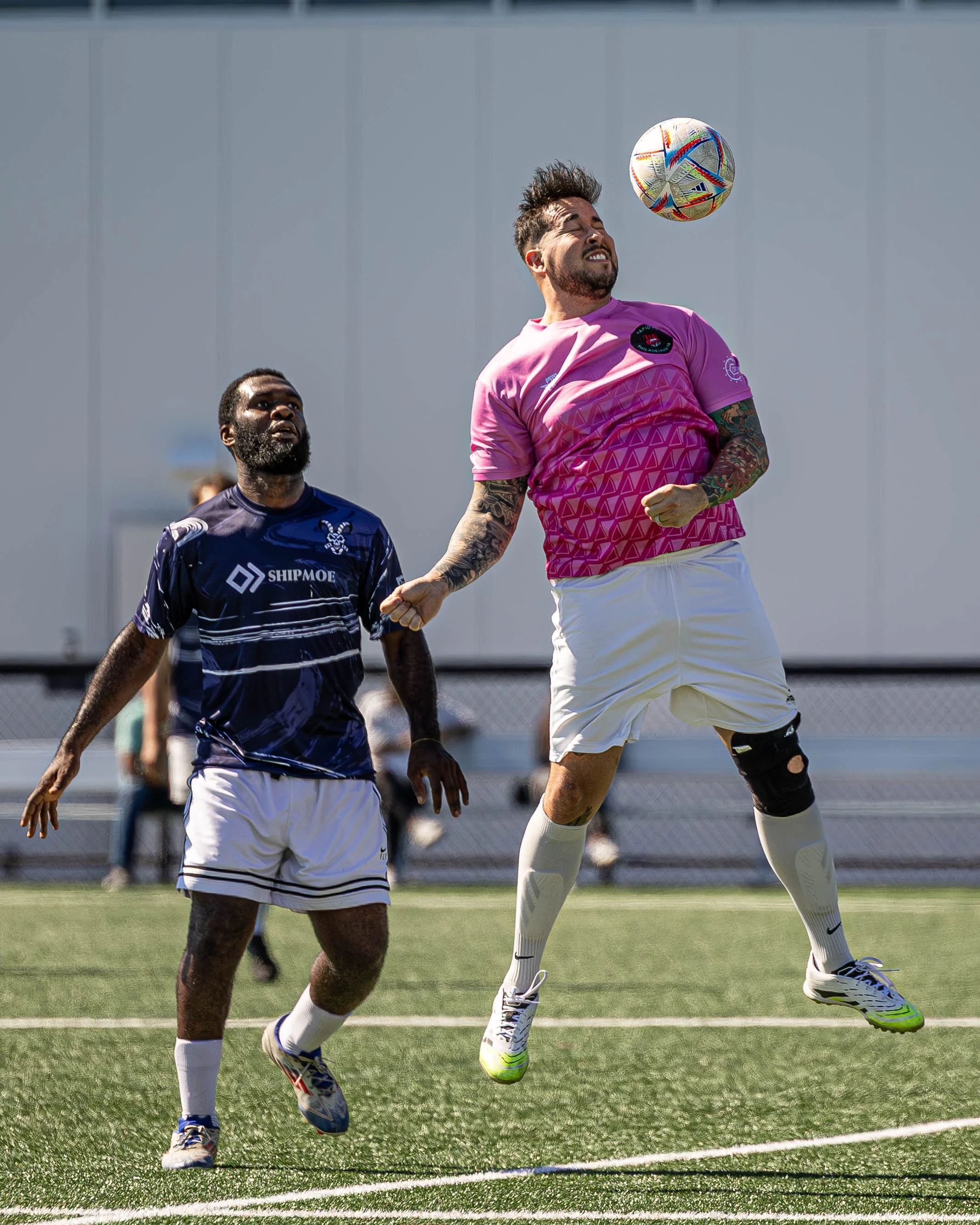 Two soccer players competing for the ball on the field, one in a pink jersey jumping to head the ball, and another in a navy jersey watching.