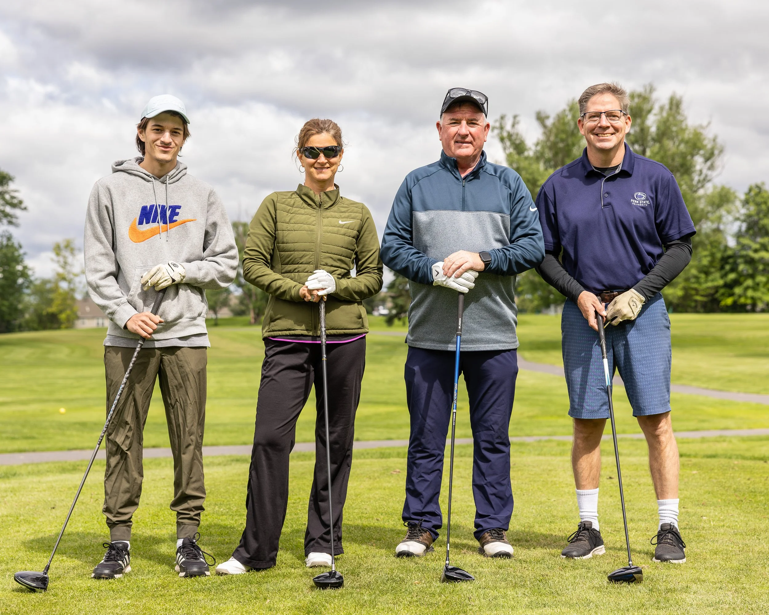 Four people standing on a golf course with golf clubs, dressed in casual golf attire, smiling, with green trees and a cloudy sky in the background.