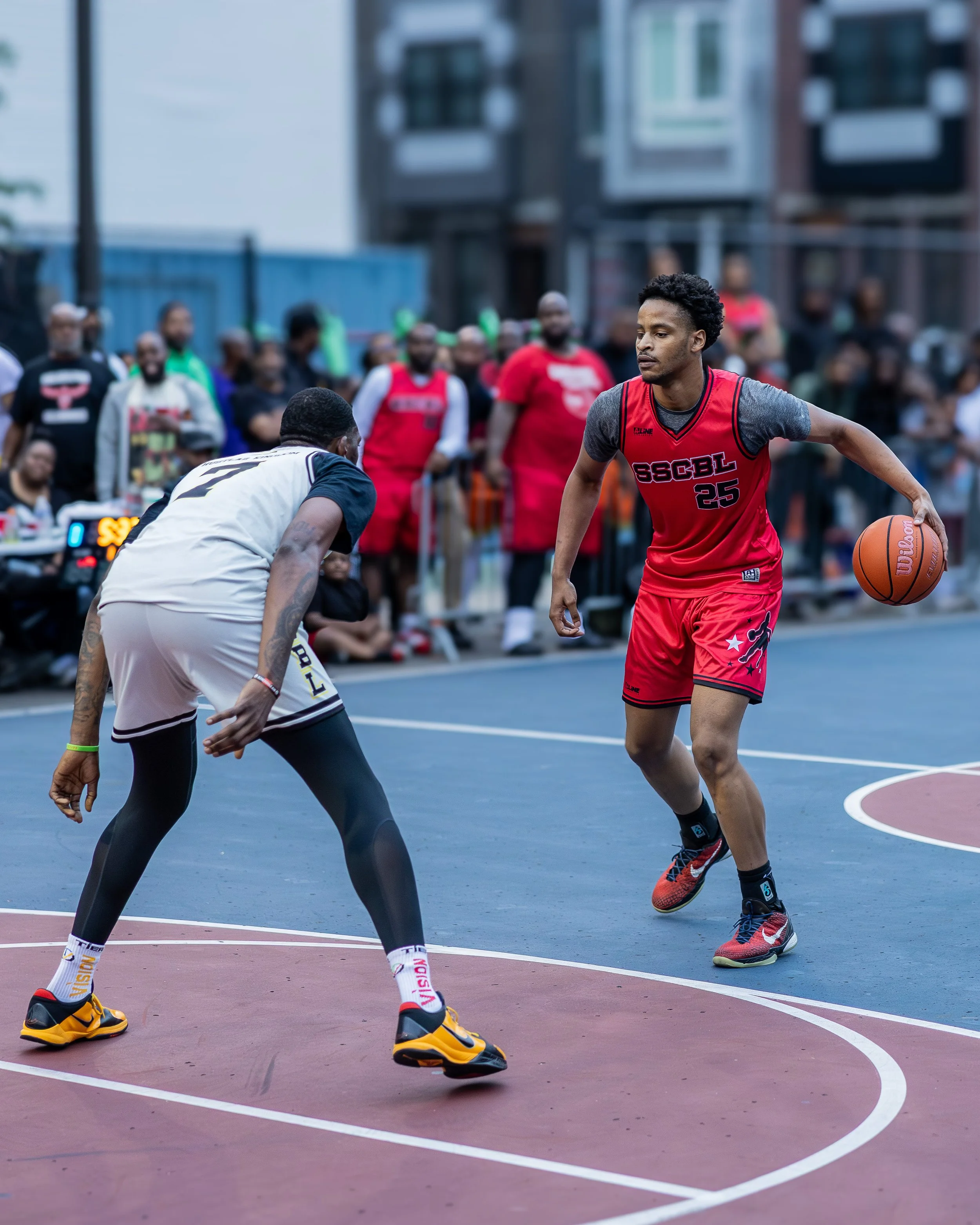 A basketball game on an outdoor court with a player in red dribbling past a defender in gray and black, with spectators watching in the background.