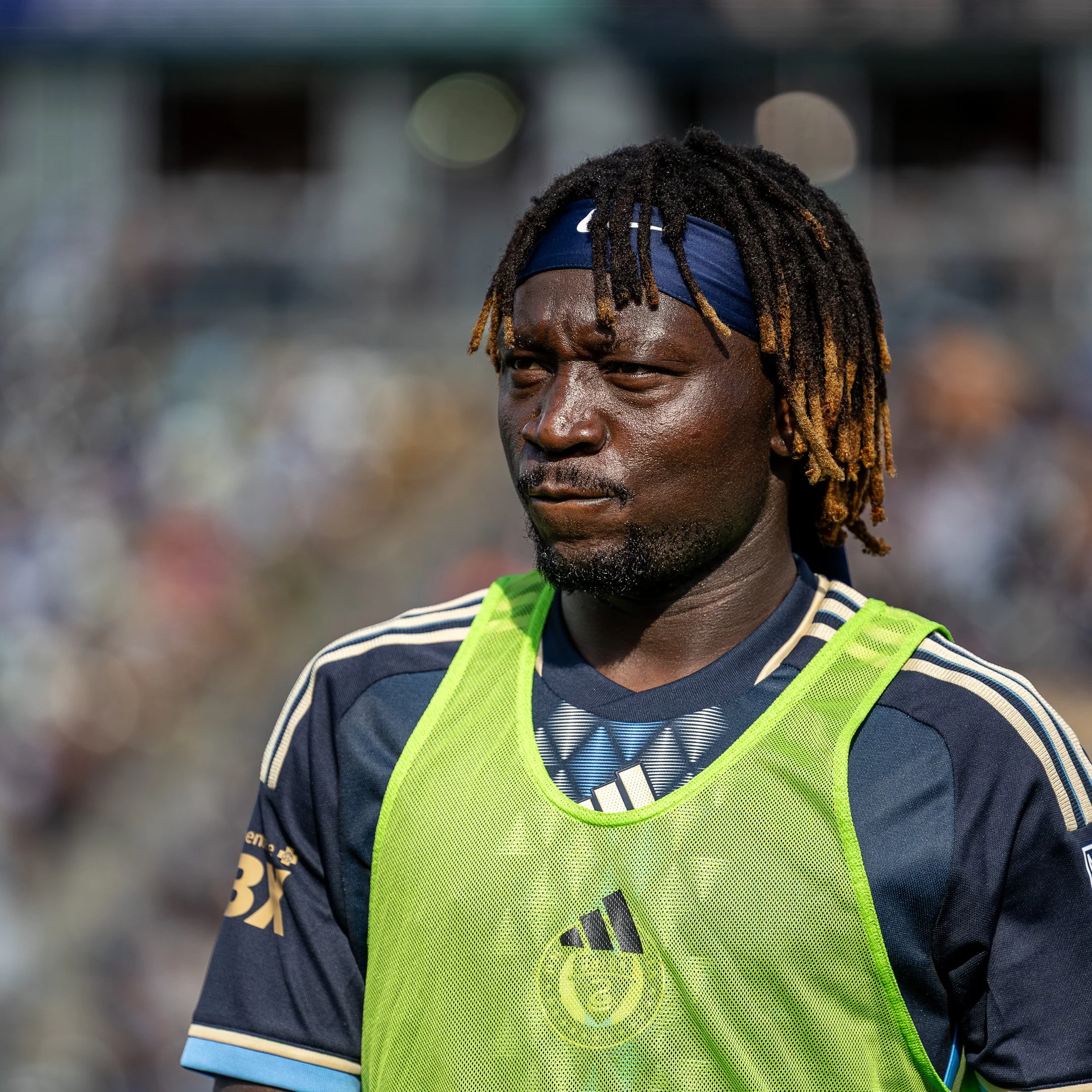 A football player with dreadlocks is wearing a blue jersey and a yellow-green training vest during a game or practice.