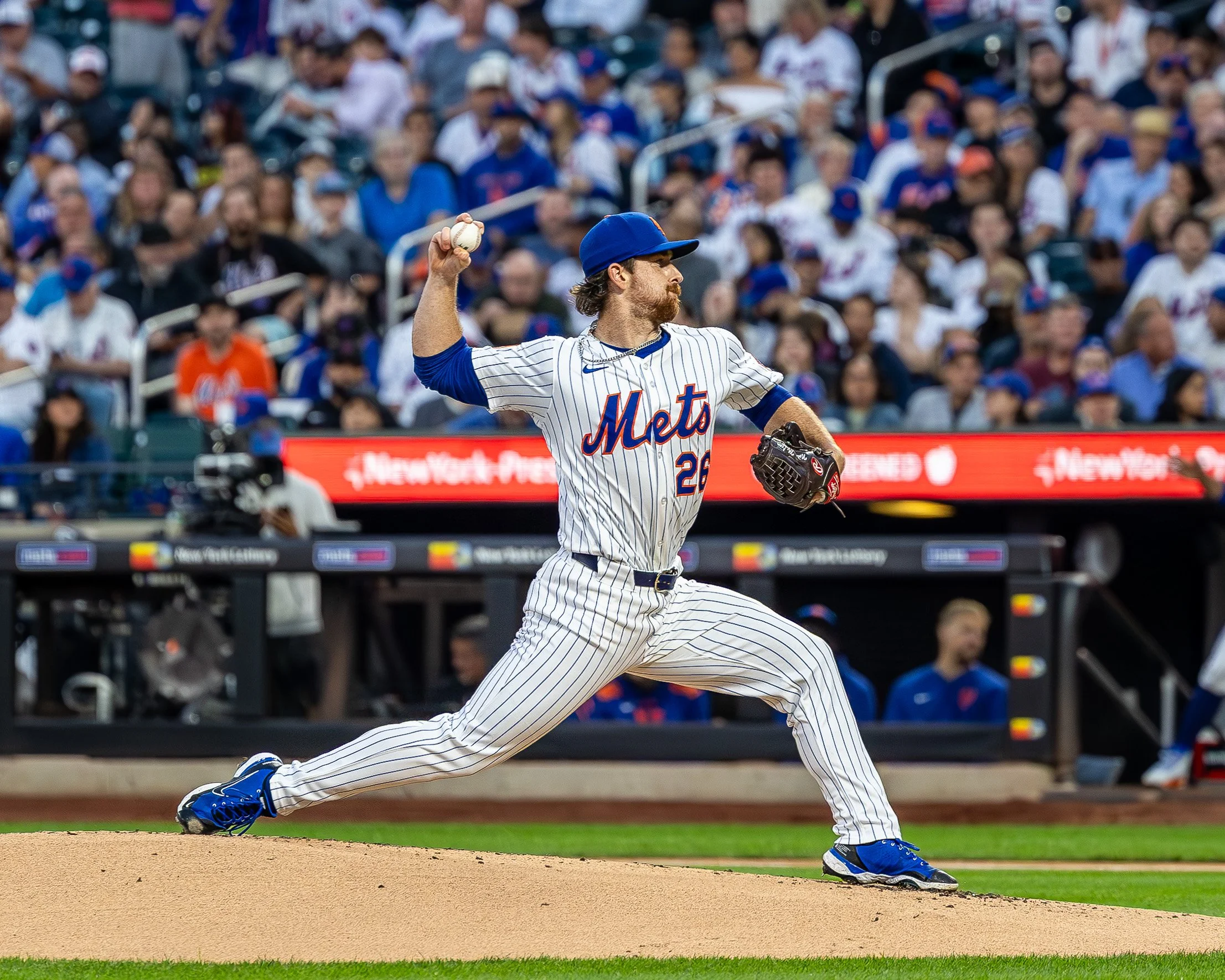 A baseball player in a New York Mets uniform throws a pitch during a game with a crowd in the background.