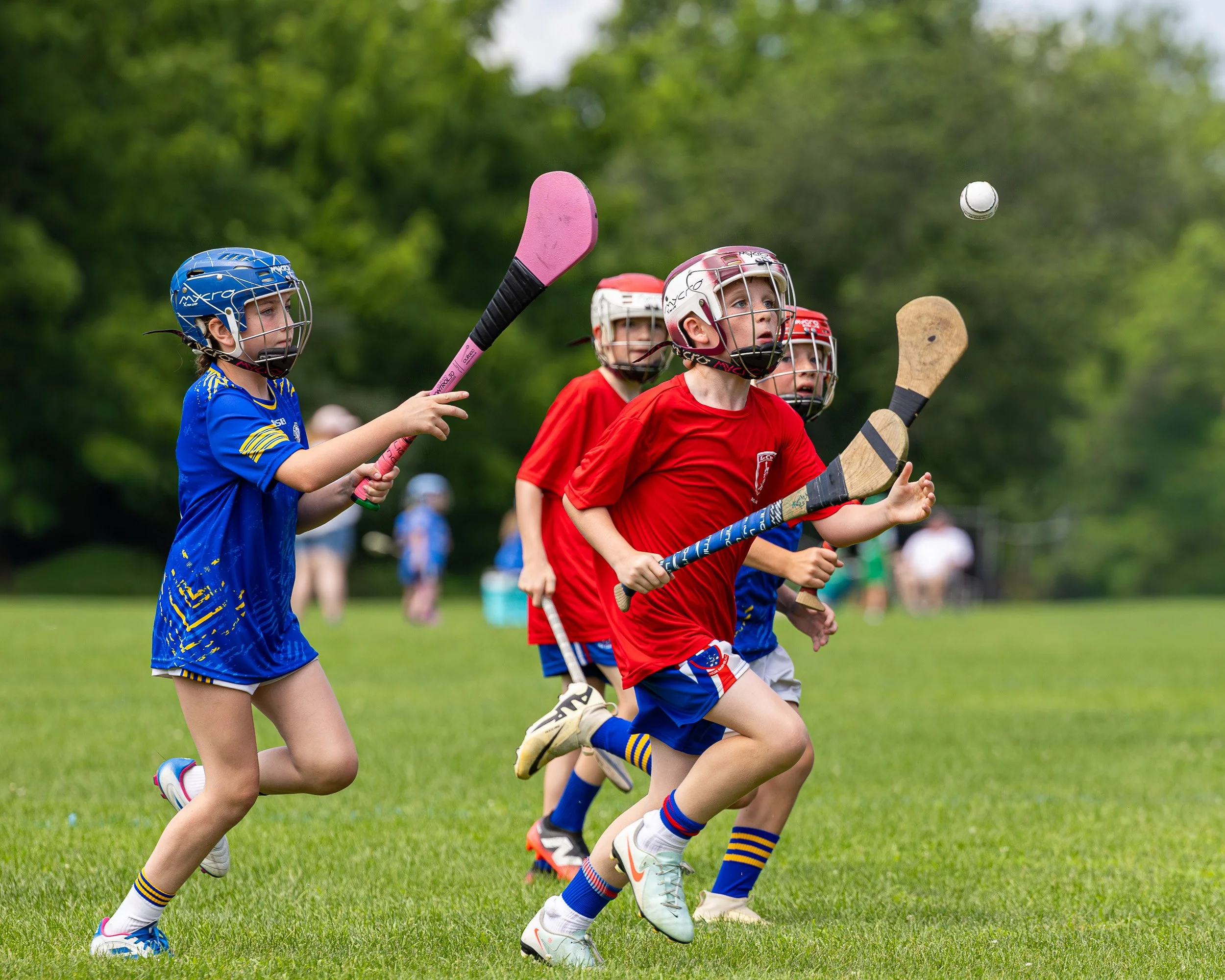 Children playing field hockey outdoors, wearing helmets and sports uniforms, actively reaching for the ball.