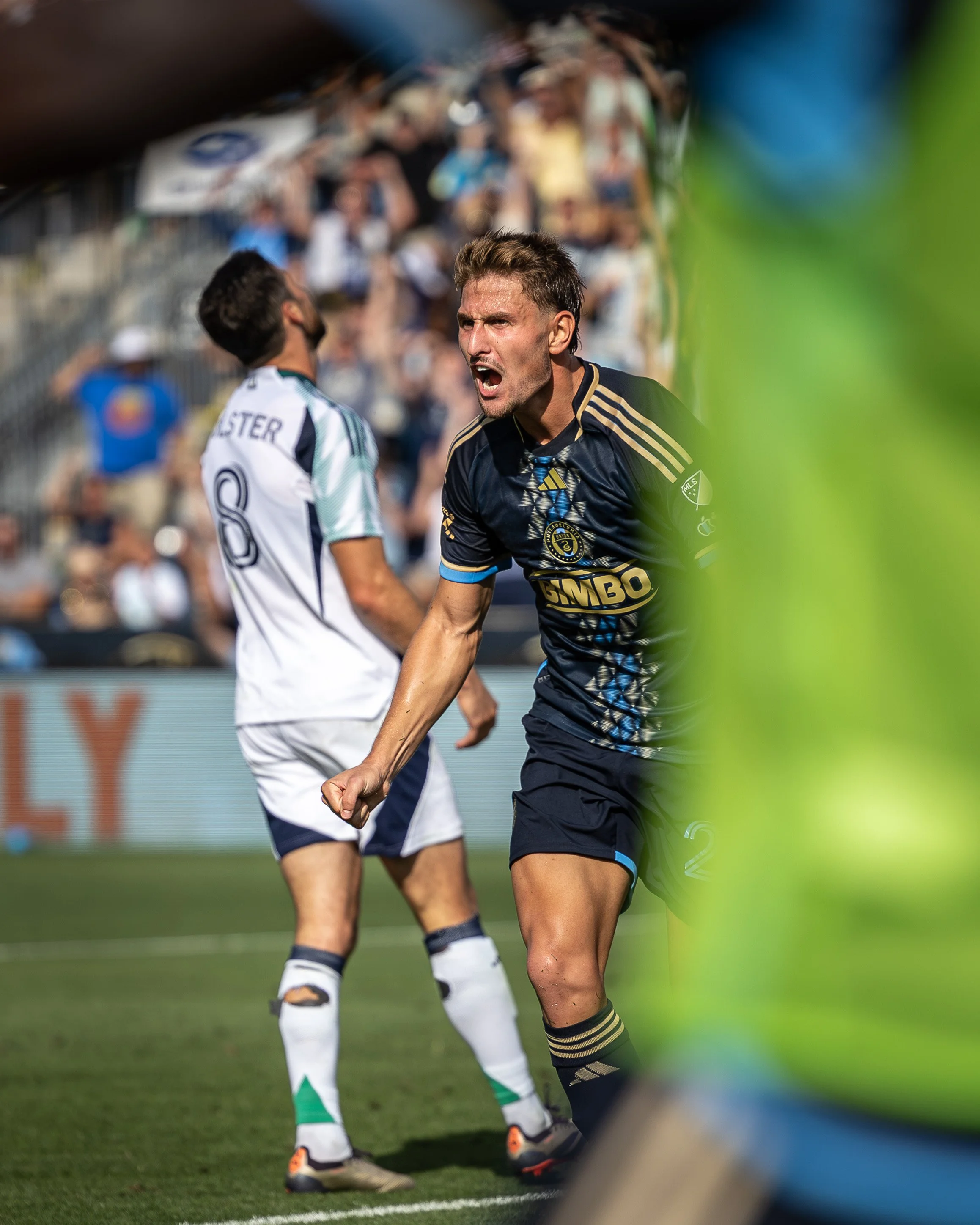 A soccer player in a navy blue jersey is celebrating with a clenched fist and an animated facial expression, next to another player in a white jersey, during a match. The scene is set in a stadium with spectators in the background.