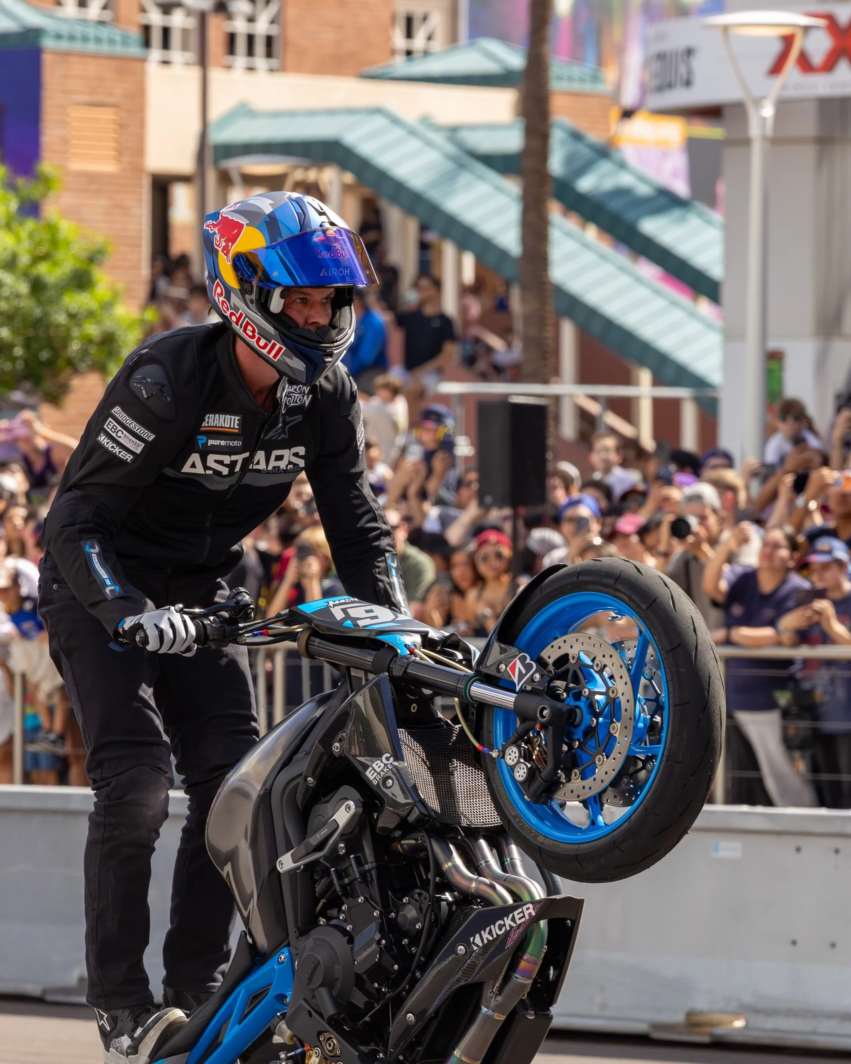 Motorcycle stunt rider performing a wheelie on a racing bike in front of a crowd at an outdoor event.