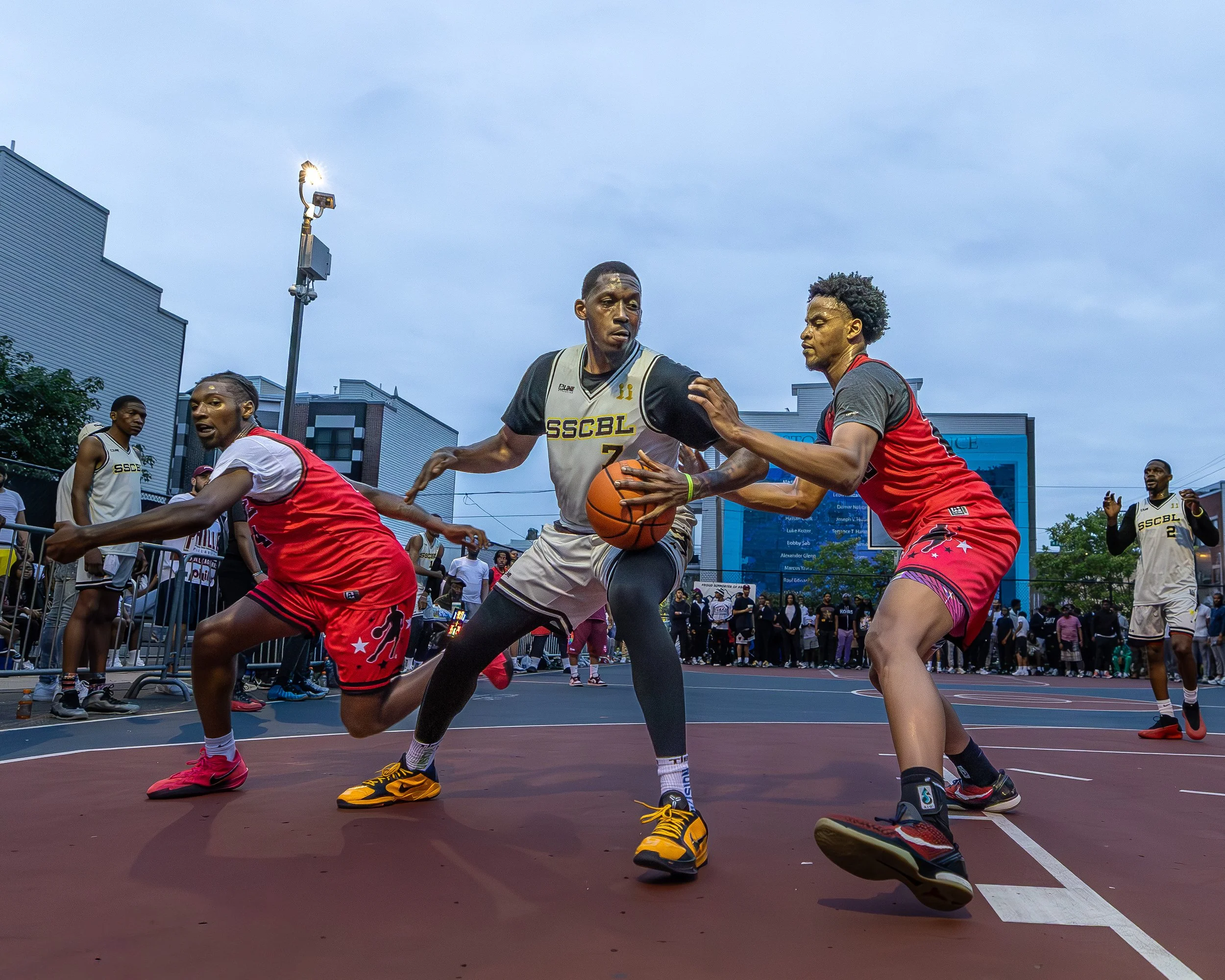 Players competing in a street basketball game on an outdoor court, with spectators watching in the background.