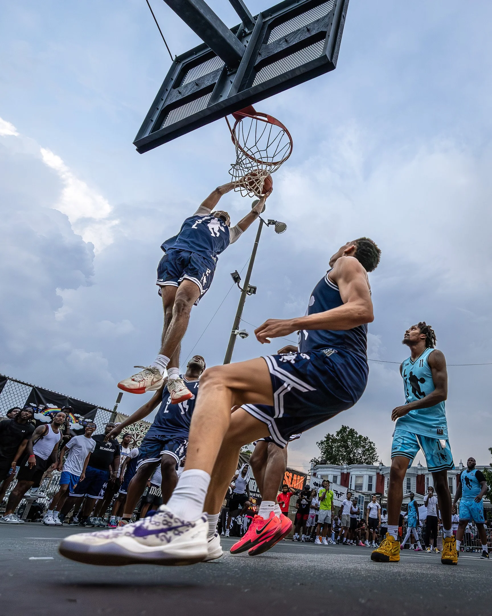 Basketball game with players jumping for a shot, crowd watching, outdoor court under cloudy sky.