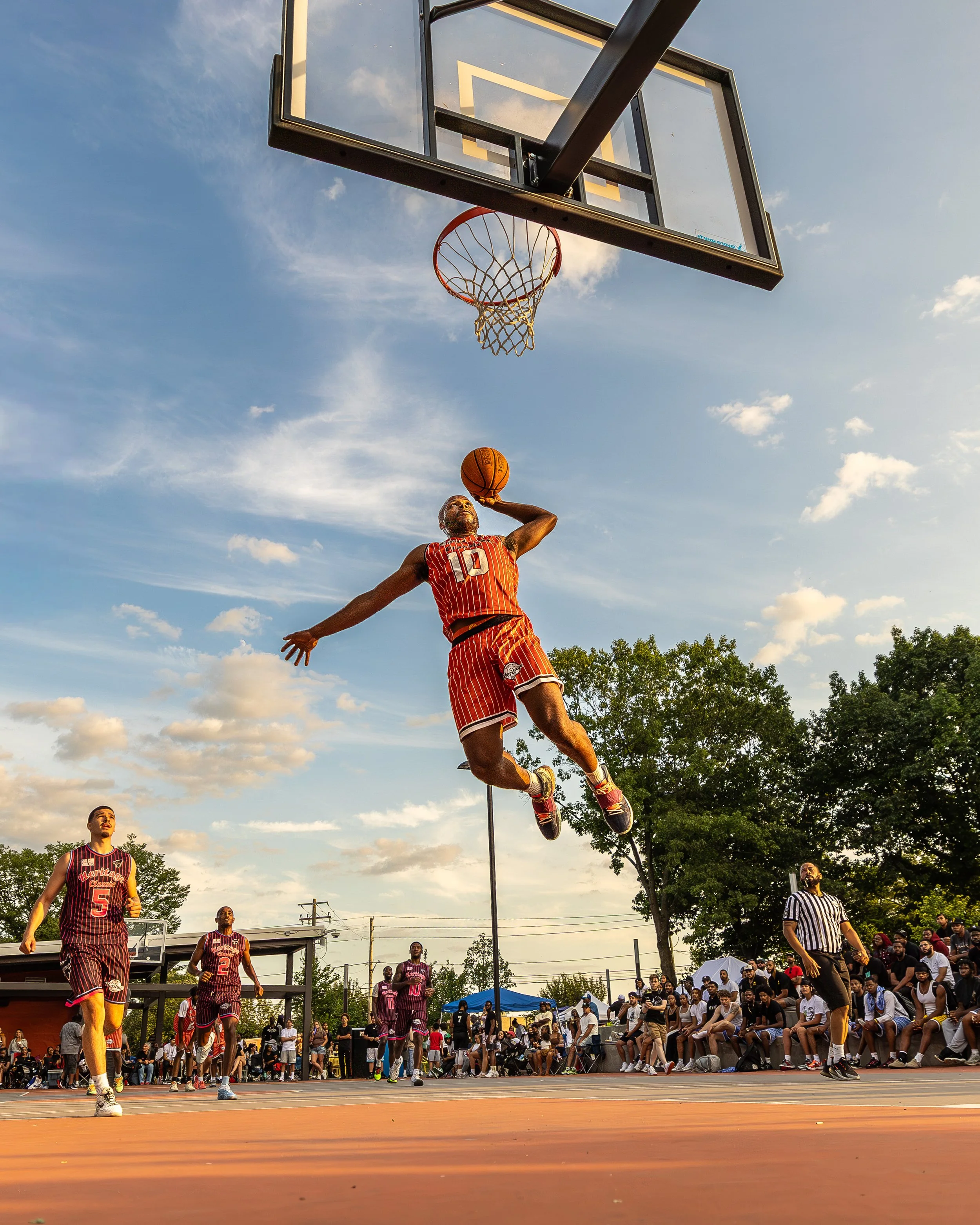 A basketball player in a red and orange striped jersey is jumping to make a shot during a game on an outdoor court, with a crowd of spectators watching.