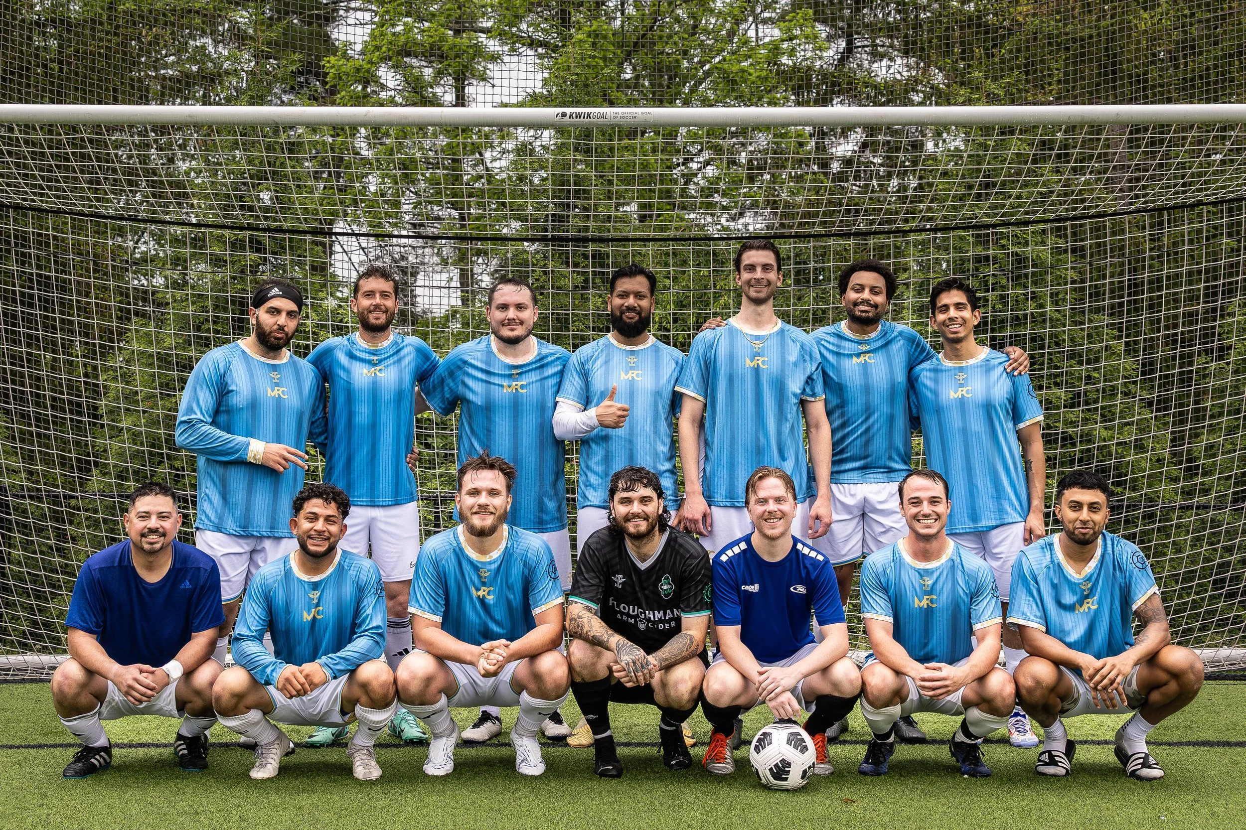 A group of 14 soccer players in blue and black jerseys posing in front of a goal on a soccer field, with trees in the background.