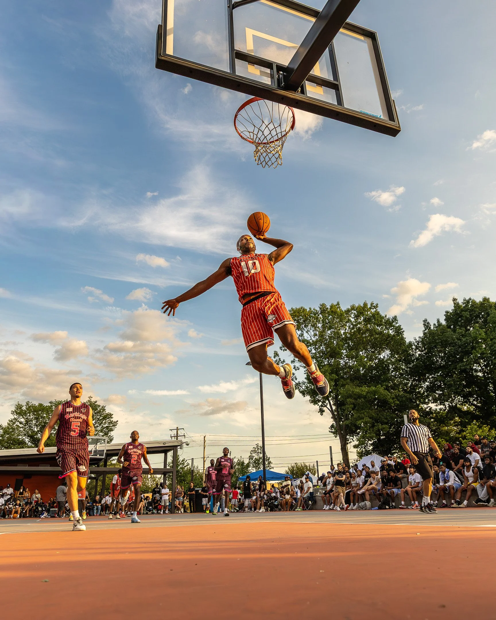 A basketball player in a red and yellow uniform leaps towards the hoop with a basketball in hand during an outdoor game, with spectators watching on and a clear sky overhead.