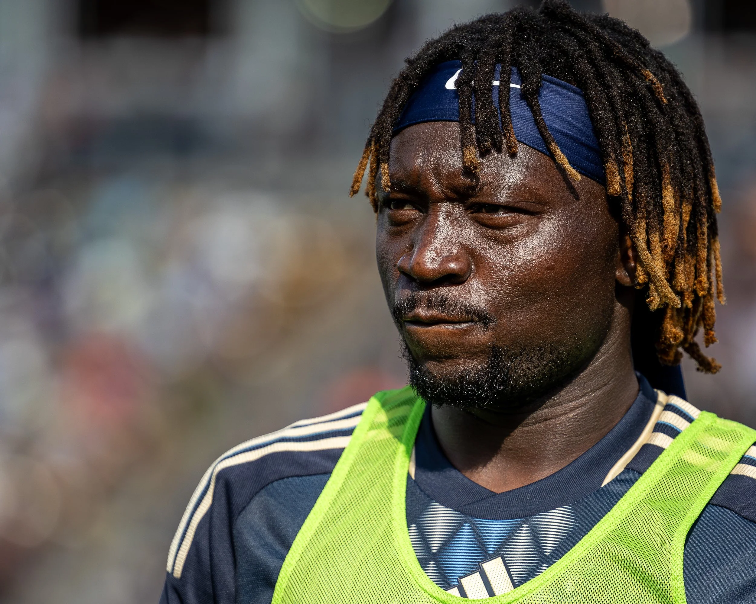 Close-up of a male athlete with dreadlocks, wearing a blue Nike headband and a neon green sports bib, on a sports field.