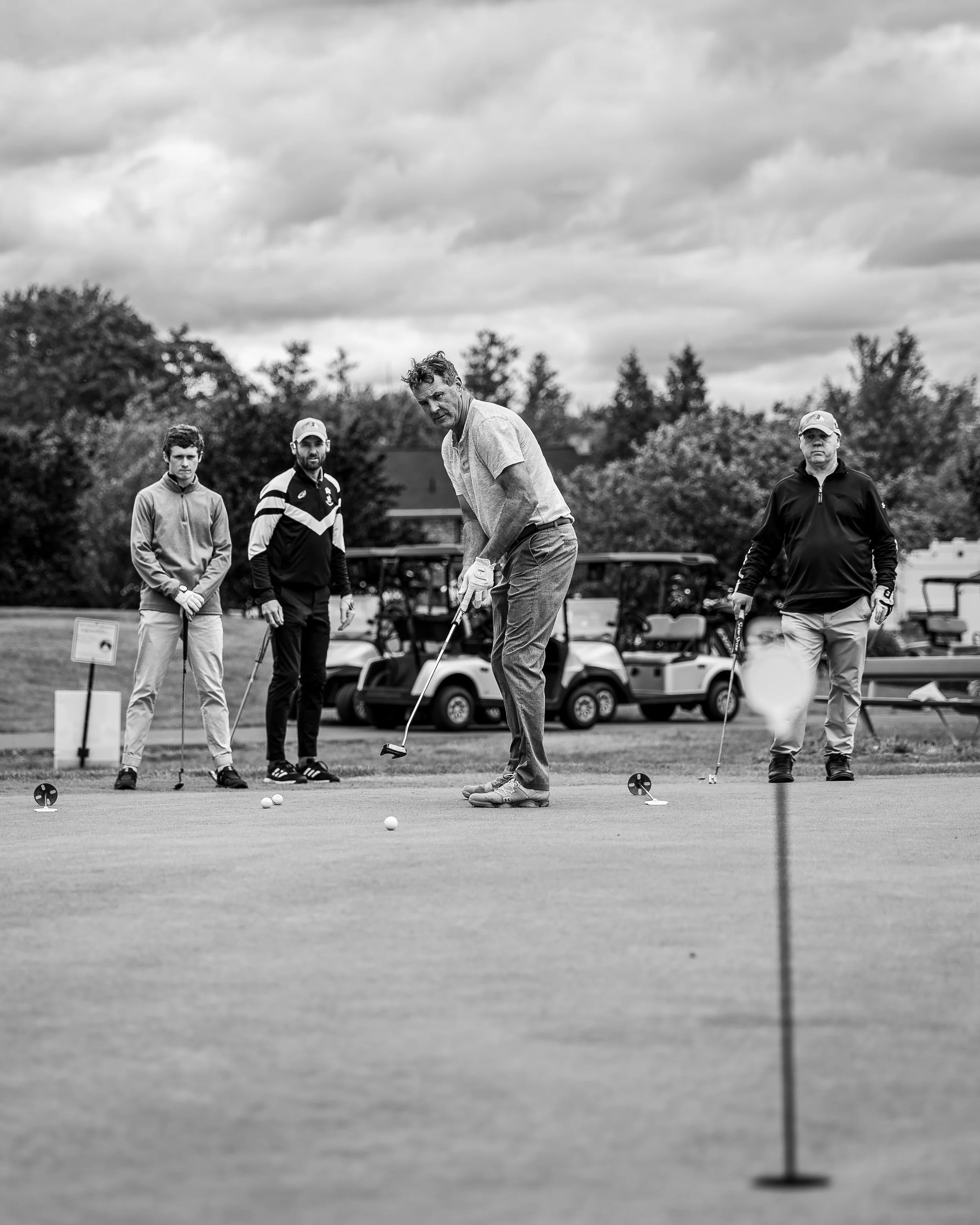 Four men playing golf on a golf course, with a golf flag in the foreground and golf carts in the background, under a cloudy sky.