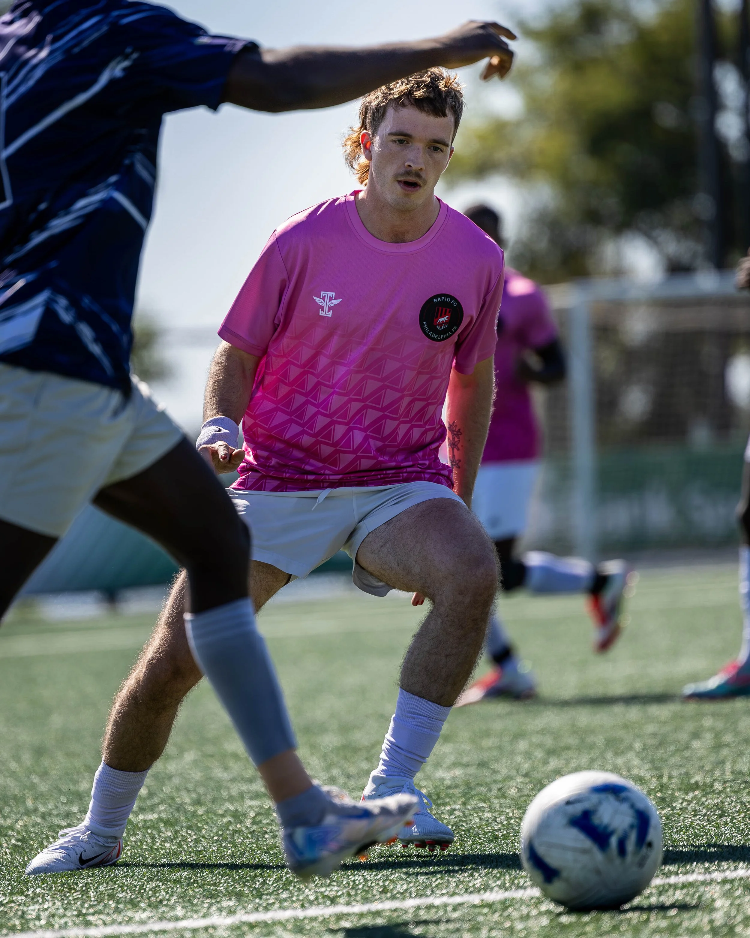 A young male soccer player in a pink jersey and white shorts is playing soccer on a field, with others in the background. He is in a low, athletic stance, focusing on the ball.