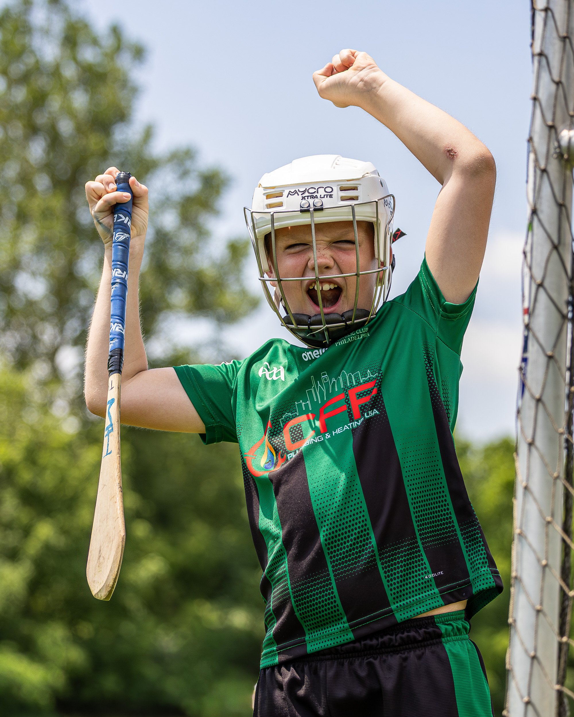 Young boy celebrating with a raised fist, wearing a white helmet and sports jersey, holding a wooden hurley stick, next to a sports goal post on a sunny day.