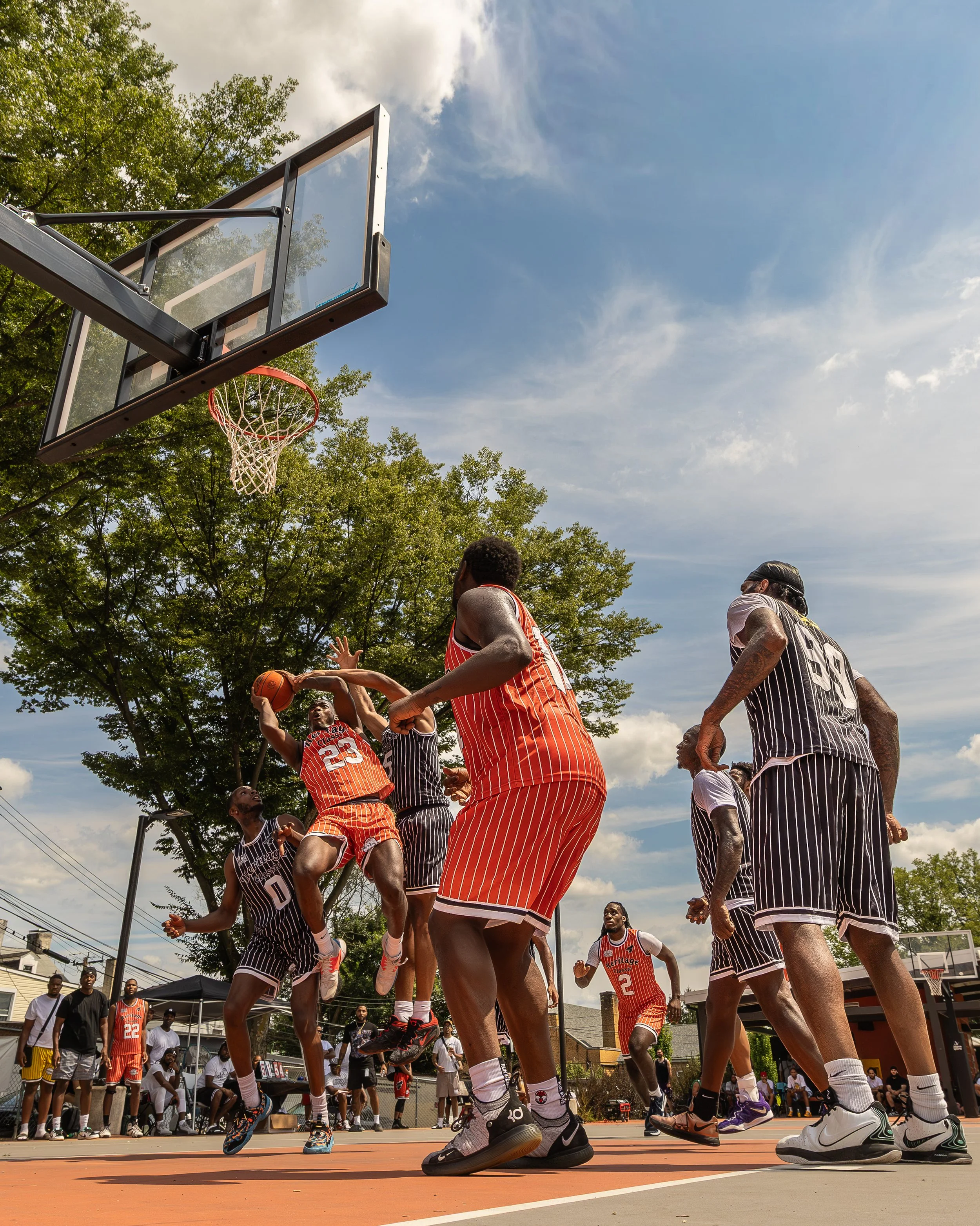 Basketball game in progress on an outdoor court with players in red and black uniforms, some jumping to shoot or block the ball. Spectators sit and stand around the court, with trees and blue sky in the background.