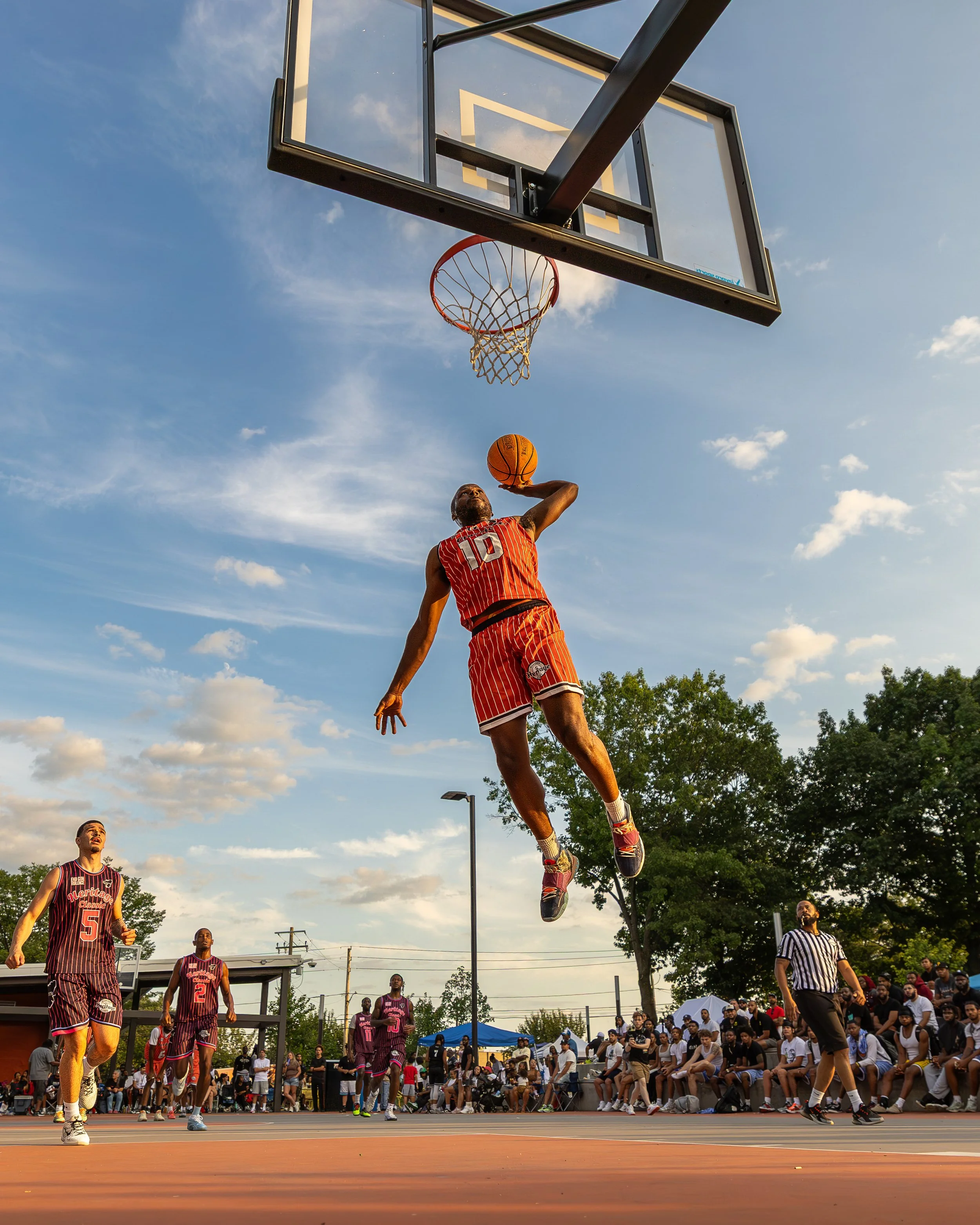 Basketball player in red uniform jumping to shoot the ball towards the hoop during a game on an outdoor court, with spectators watching in the background.