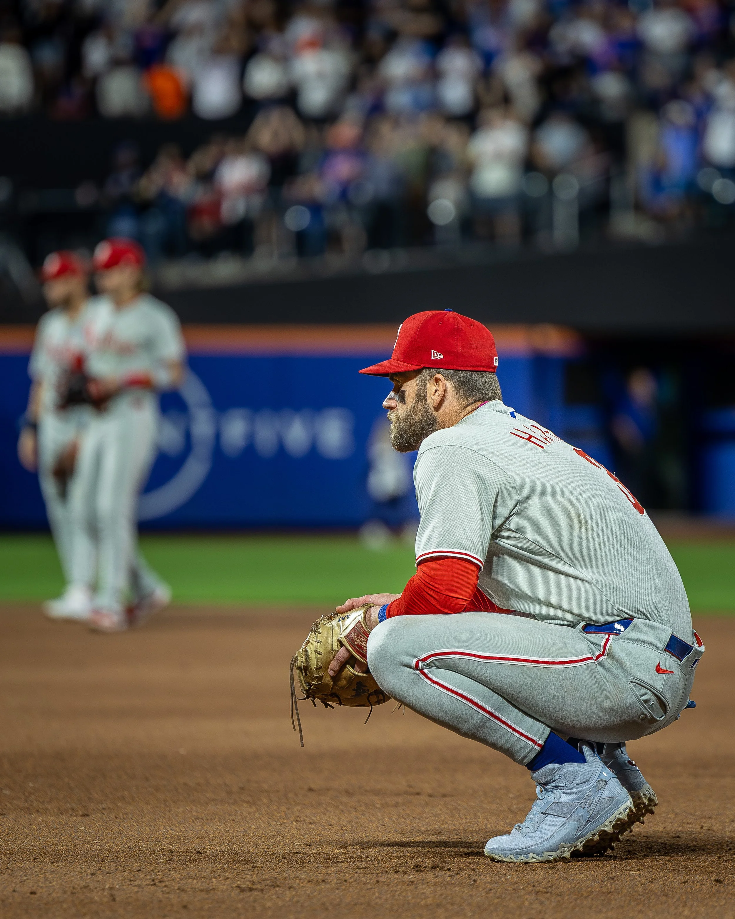 A baseball player squatting on the field in a gray uniform with red accents, wearing a red cap and cleats, with two other players in the background and a crowd in the stands.