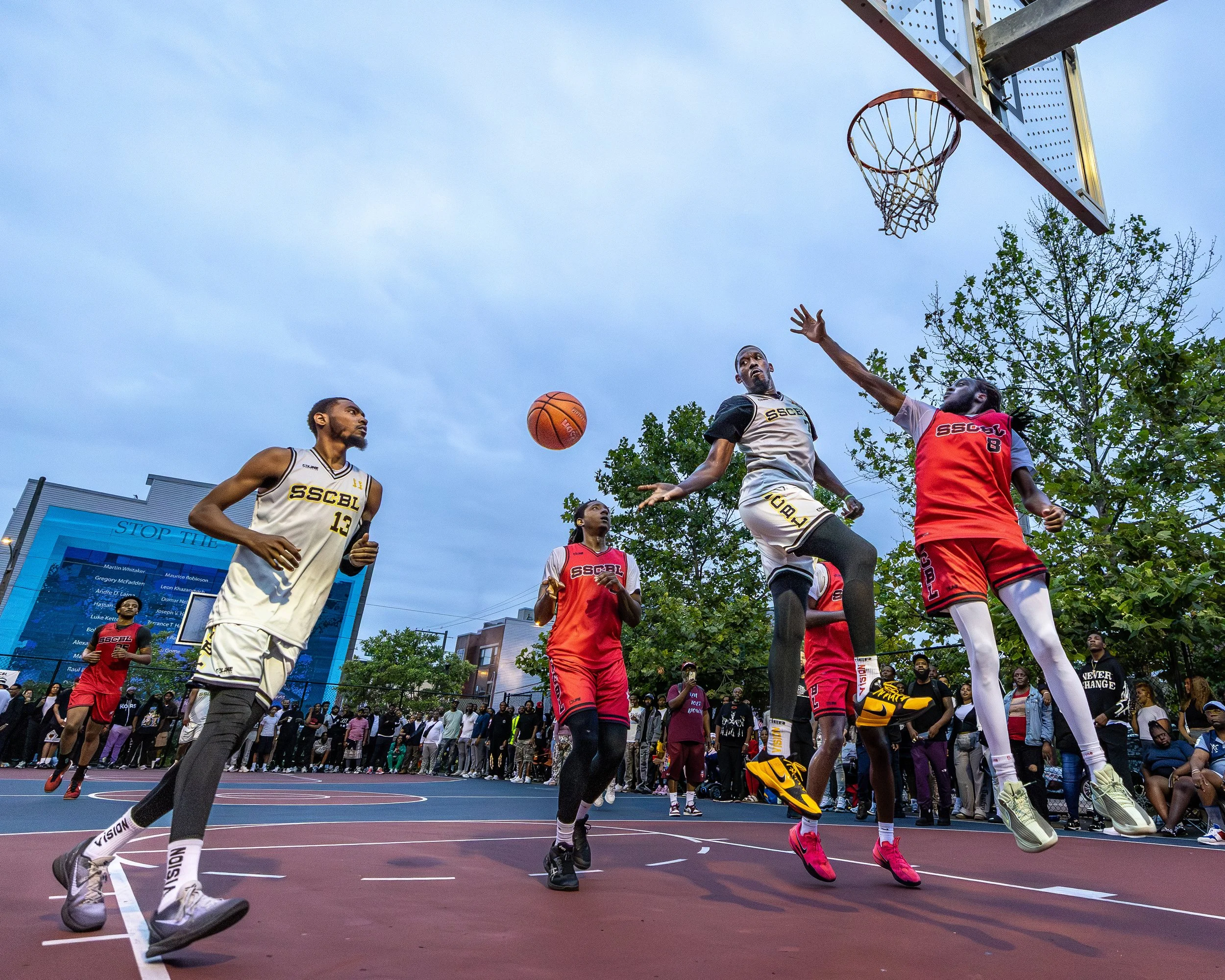 A basketball game on an outdoor court with players jumping to contest a shot, surrounded by spectators and trees, under a cloudy sky.