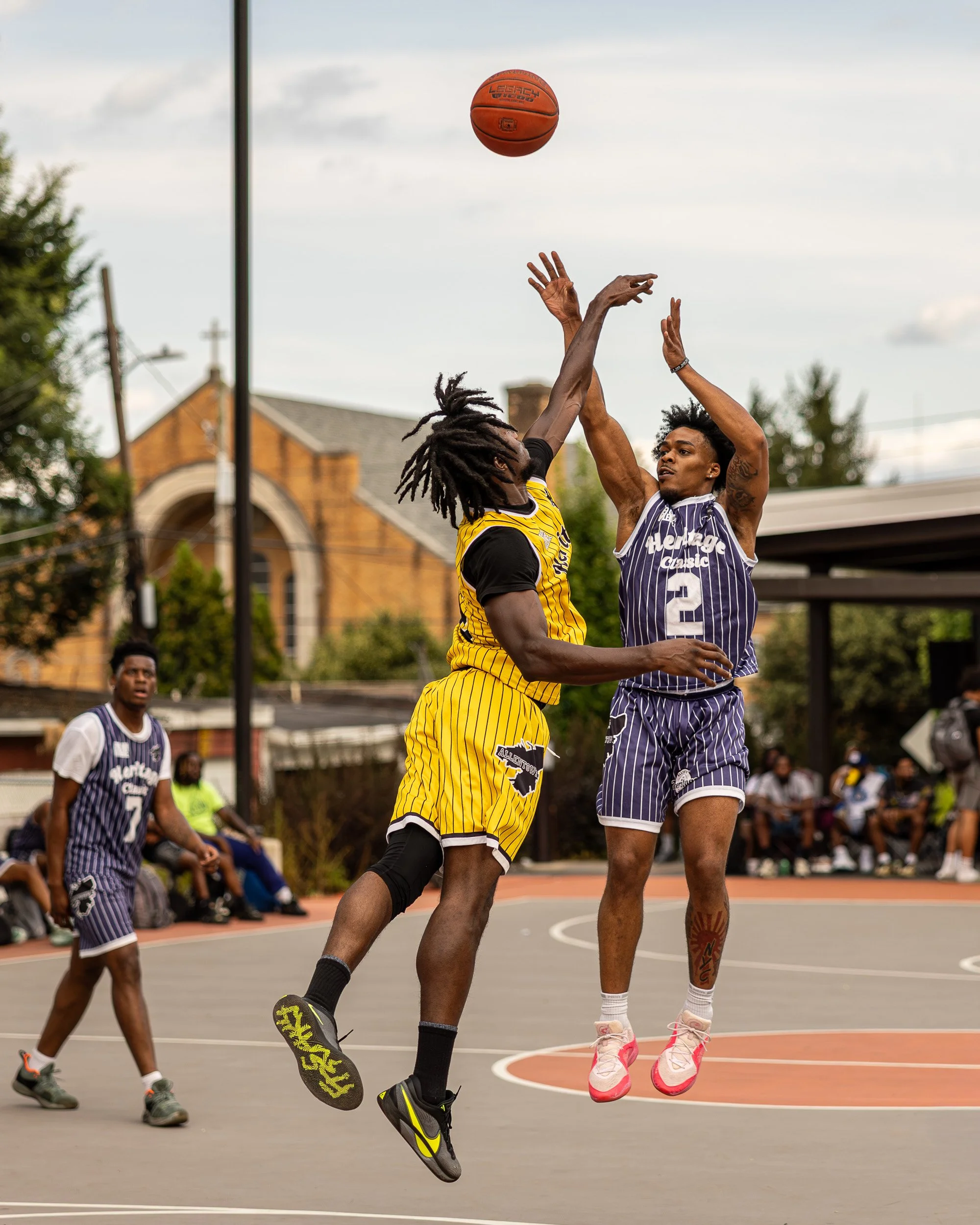 Two basketball players are jumping to catch or contest a basketball in mid-air during a game. One player wears a yellow jersey with black stripes, and the other wears a blue jersey with white stripes. The game is outdoors, with spectators sitting on 