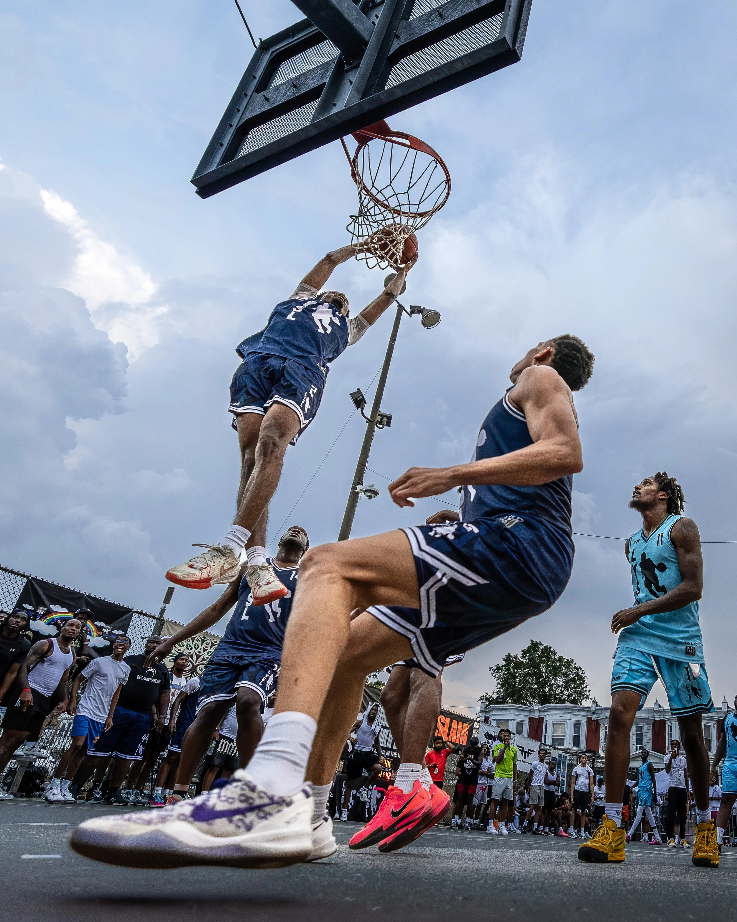 A basketball player in a blue jersey is jumping to make a shot while another player in a blue jersey is on the ground watching. Several spectators and players are gathered around on an outdoor court, with a cloudy sky overhead.