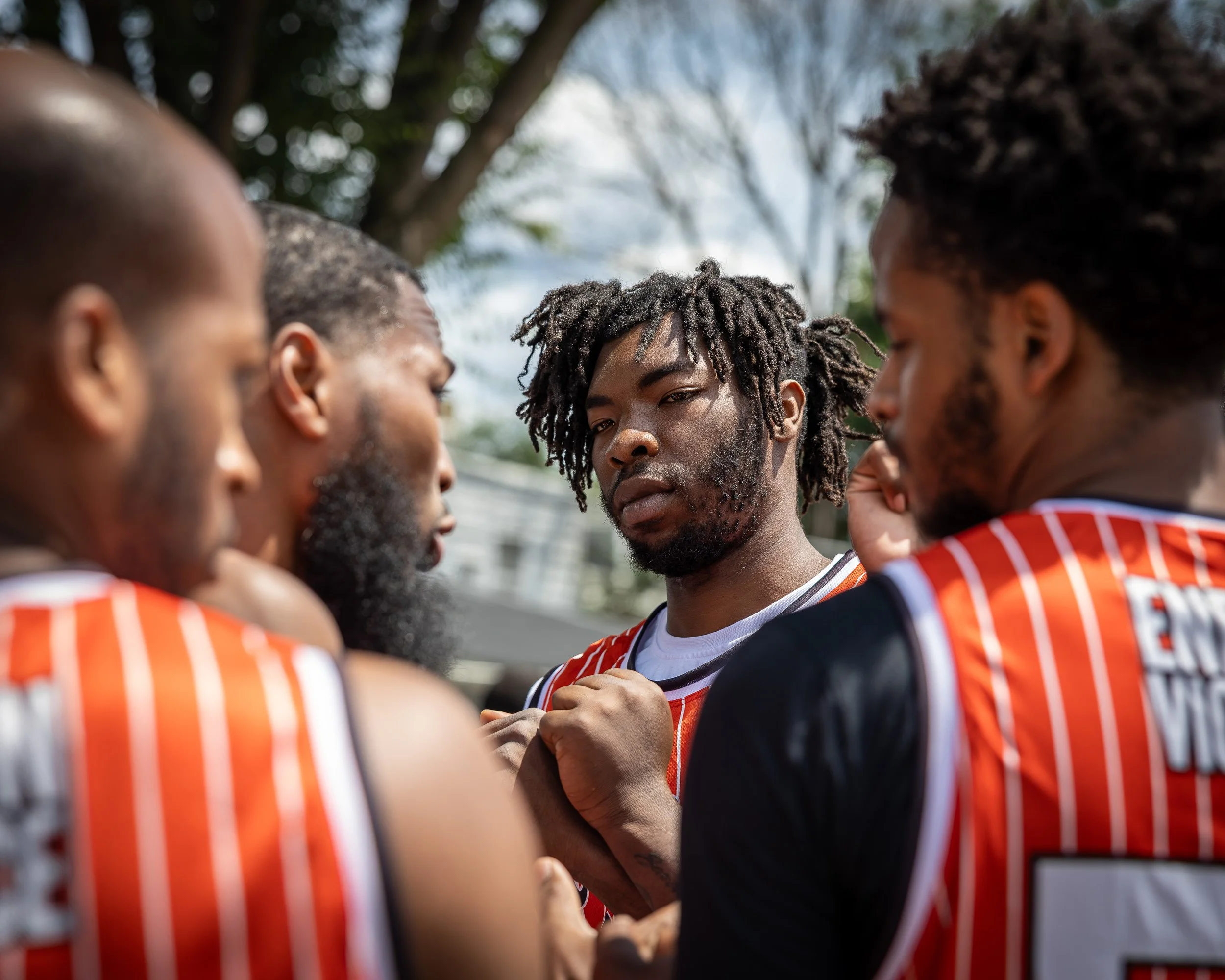 A group of young male basketball players have a team huddle outdoors, wearing orange and black jerseys, with one player in the center making a serious expression.