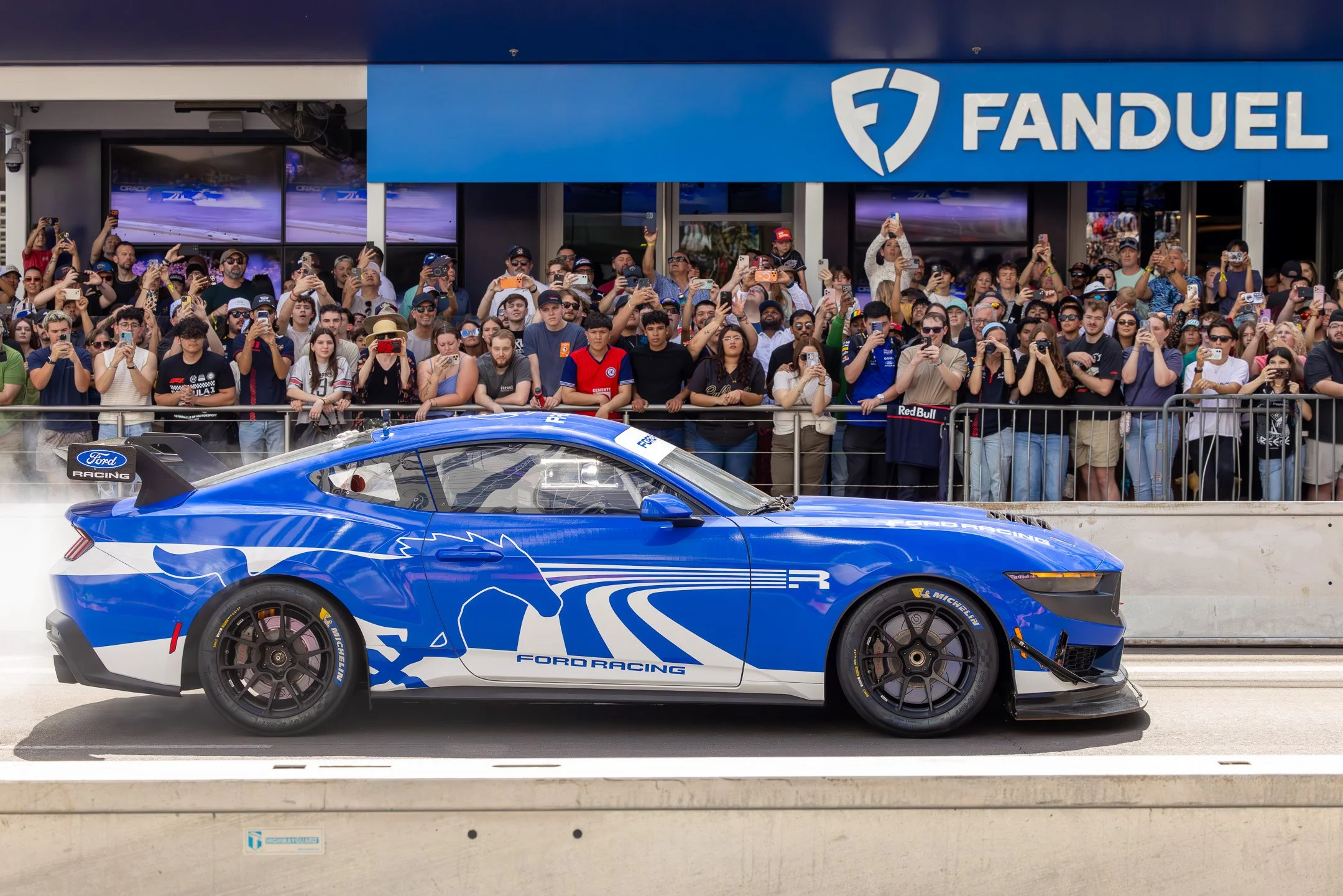 A blue Ford racing car on a racetrack with a crowd of spectators behind a barrier, taking photos, in front of a FanDuel sign.