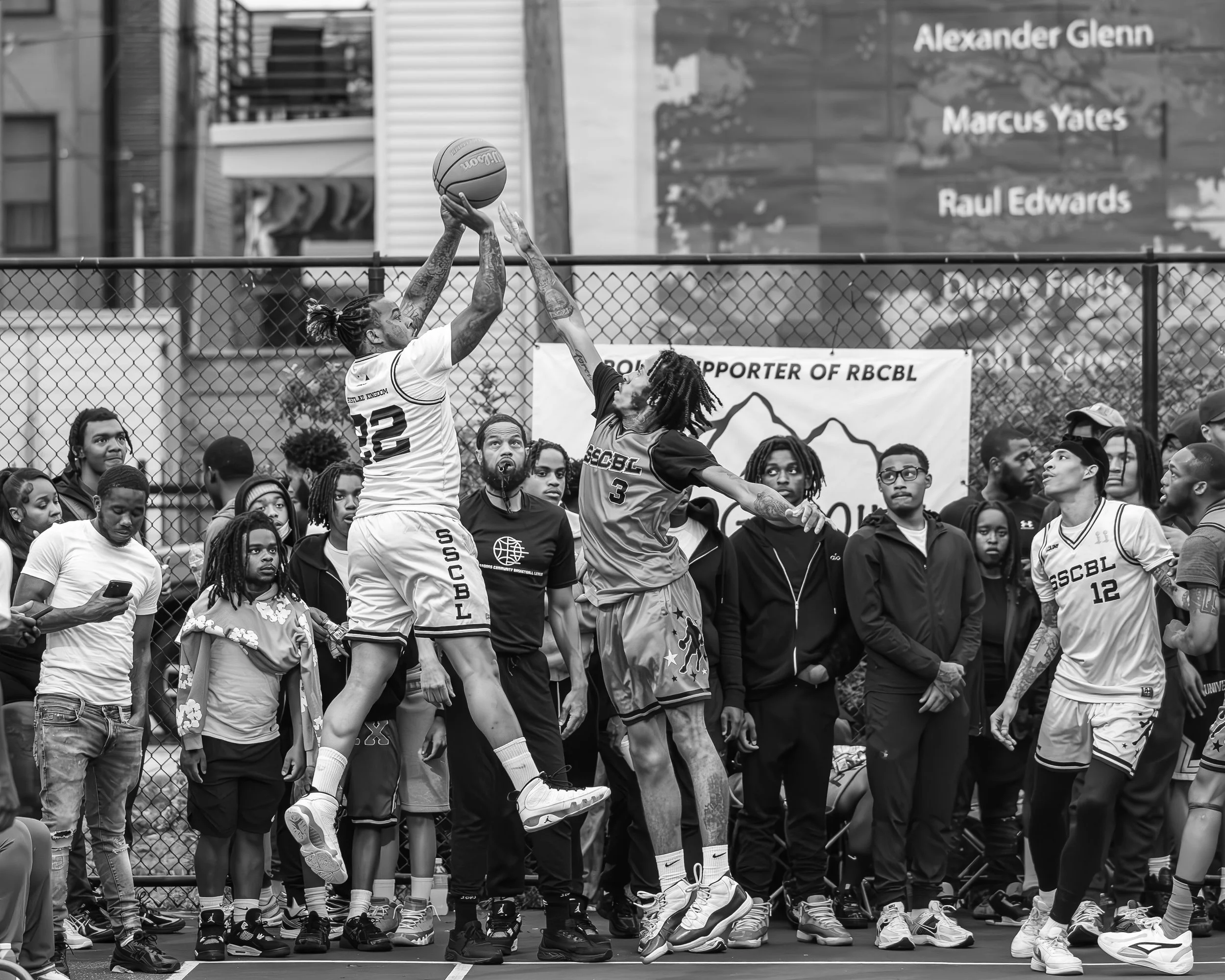 Black and white photo of two basketball players jumping for a rebound or shot during a game, surrounded by spectators and teammates on the sidelines.
