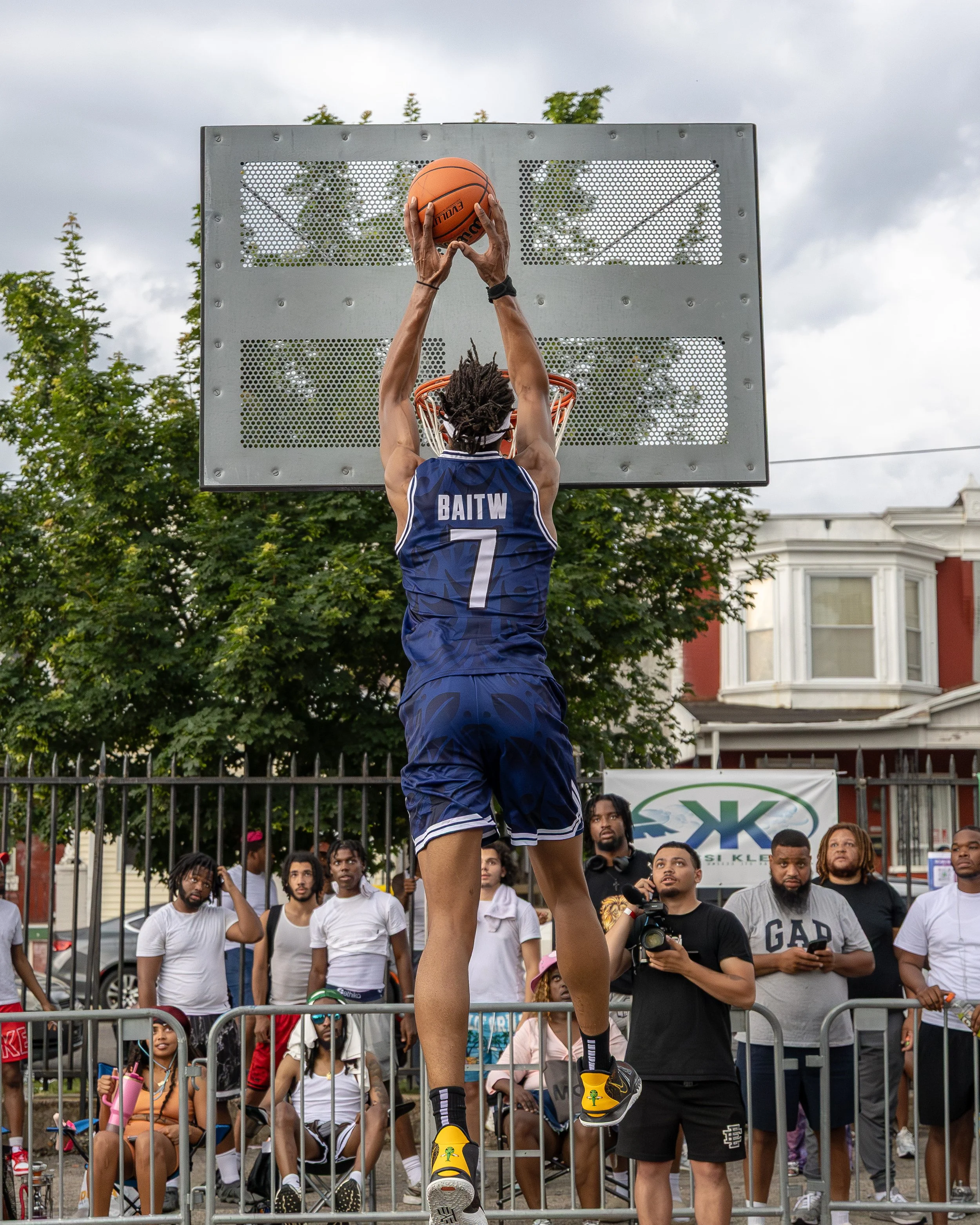 A basketball player wearing a blue jersey with the name 'Baitw' and number 7 is jumping to make a shot at the basketball hoop during an outdoor game. Several spectators and photographers watch behind a fence.