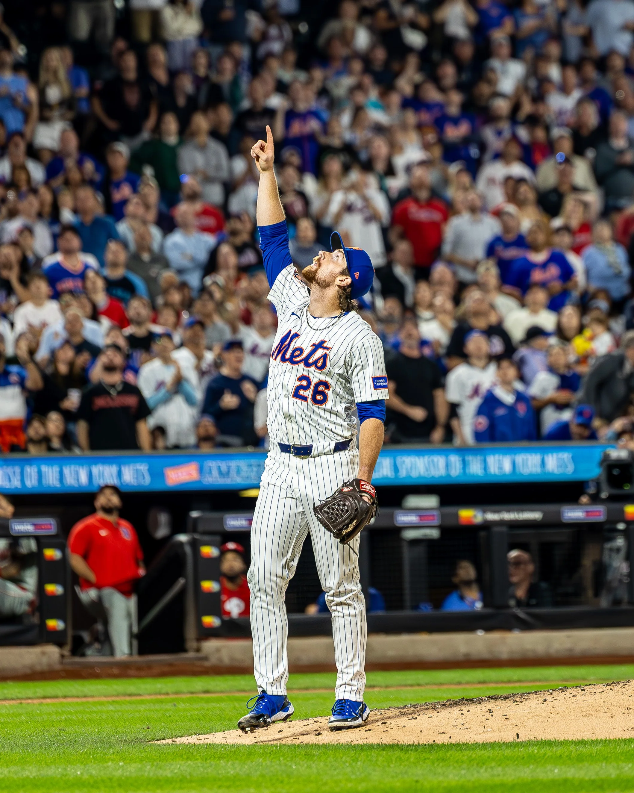 A baseball player in a pinstripe uniform with 'Mets' and the number 26, standing on the pitcher's mound, pointing upwards with his right hand, surrounded by a crowd in a stadium.