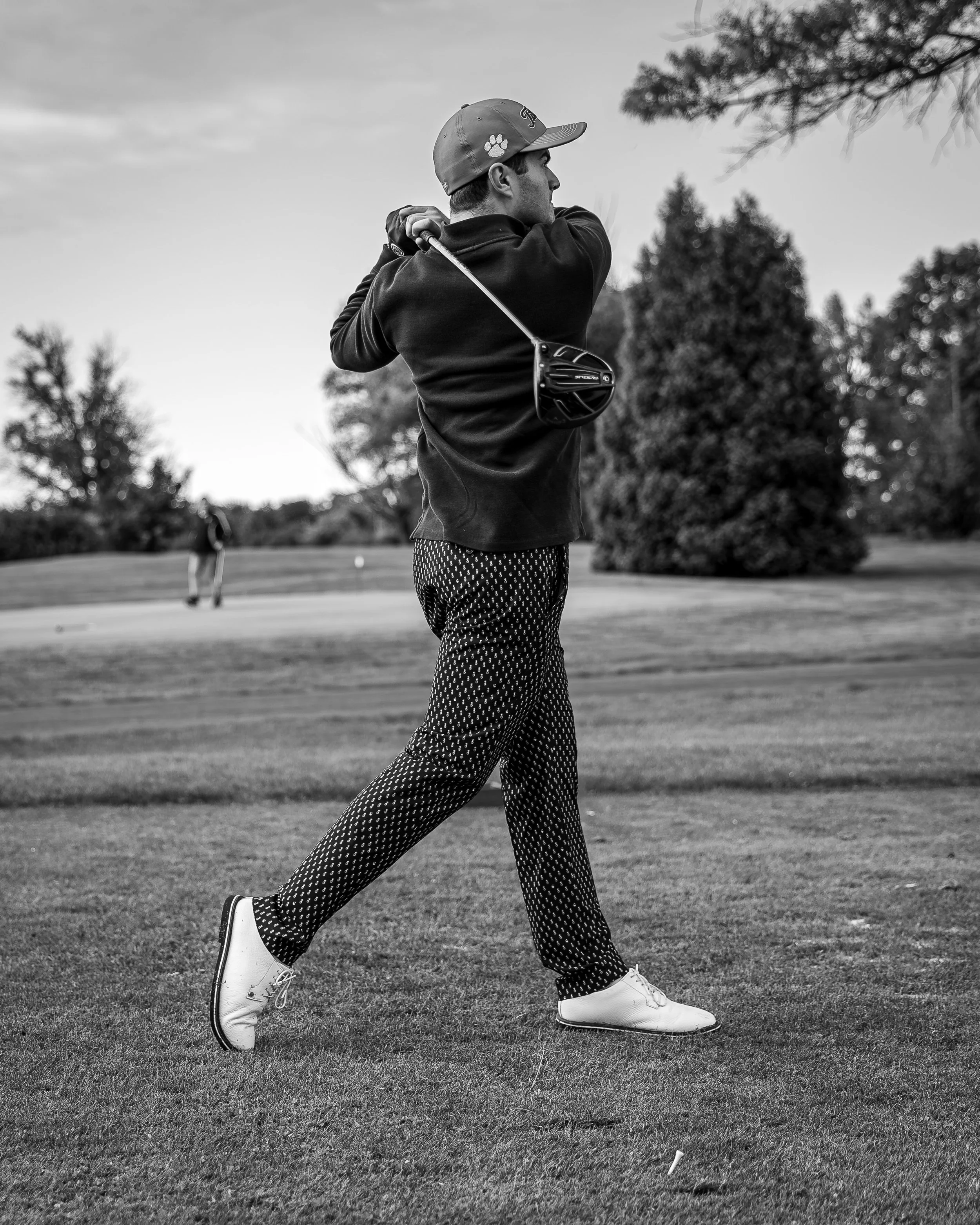 A man playing golf on a golf course, swinging a club, wearing patterned pants, a dark jacket, and a cap, with trees and another golfer in the background.