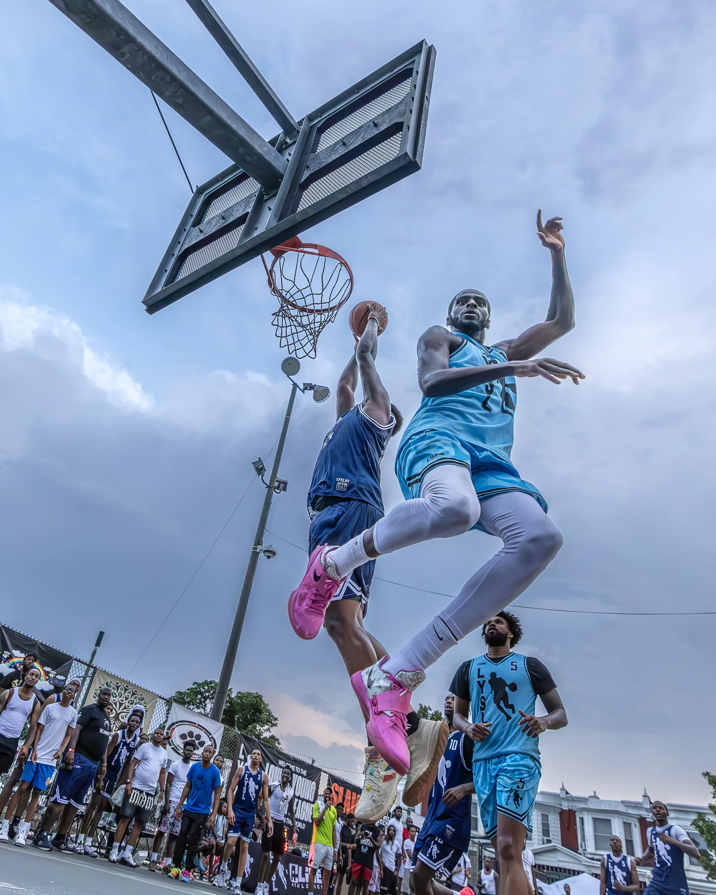 Two basketball players in blue uniforms leap towards the hoop; one is attempting a shot while the other defends. A crowd of spectators and other players watches on the court beneath a cloudy sky.