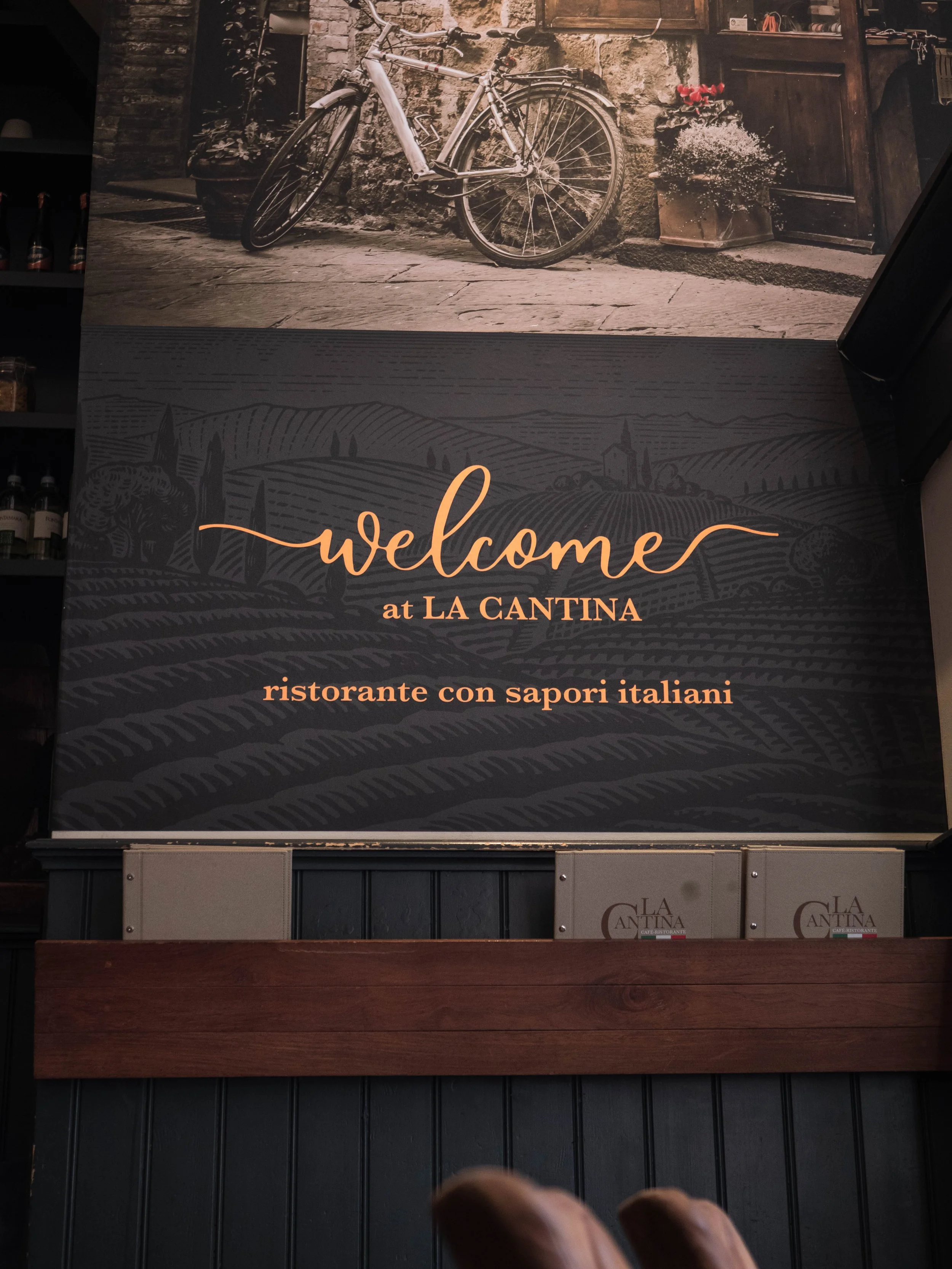 Pancarta con mensaje de bienvenida en español que dice: "welcome at LA CANTINA ristorante con sapori italiani", con fondo de paisaje de campos y colinas y arriba una foto de una bicicleta apoyada en una pared de piedra con flores