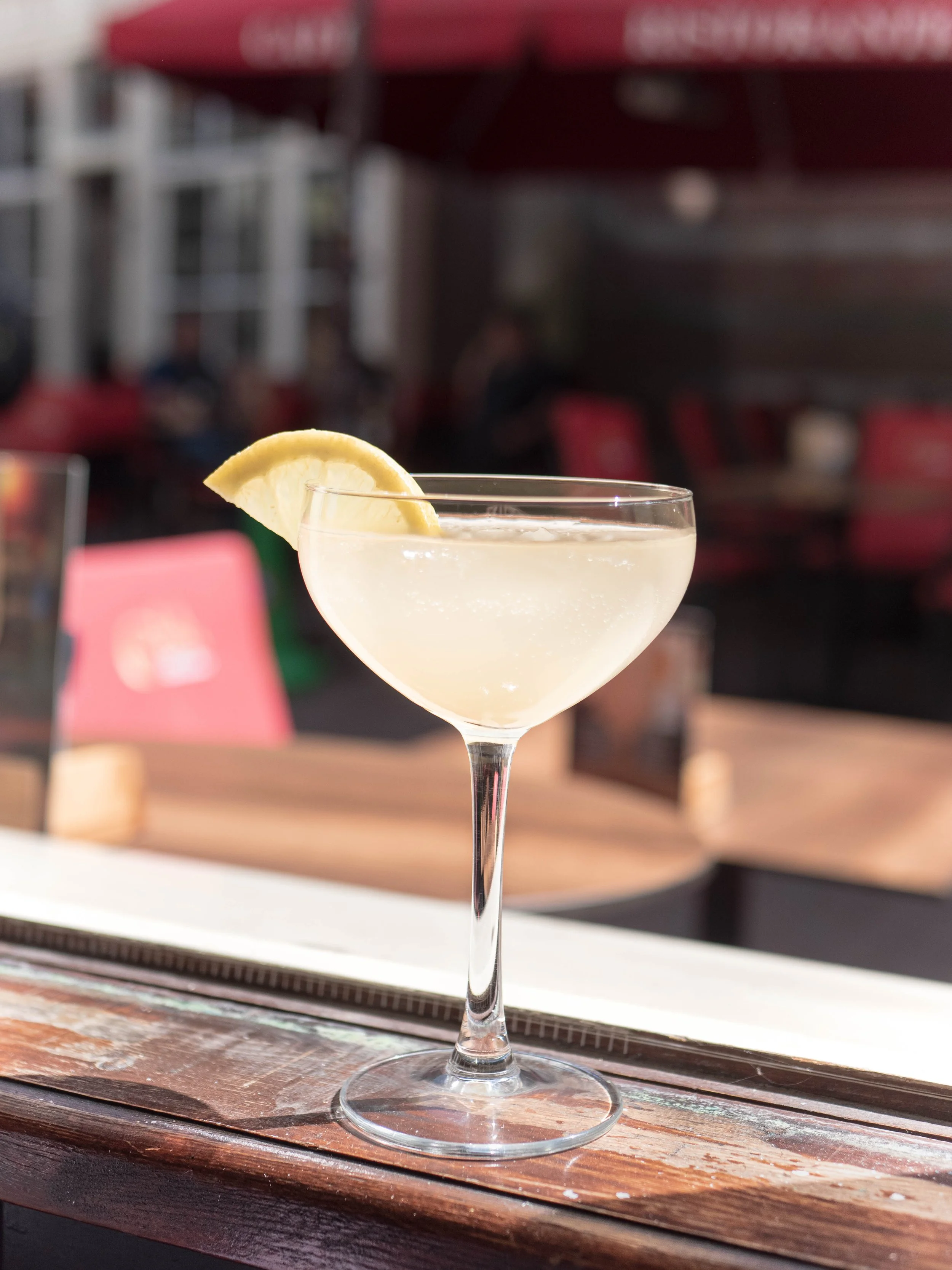 Cóctel en copa de cristal con limón como decoración, en una mesa de madera en un espacio de restaurante o bar al aire libre.
